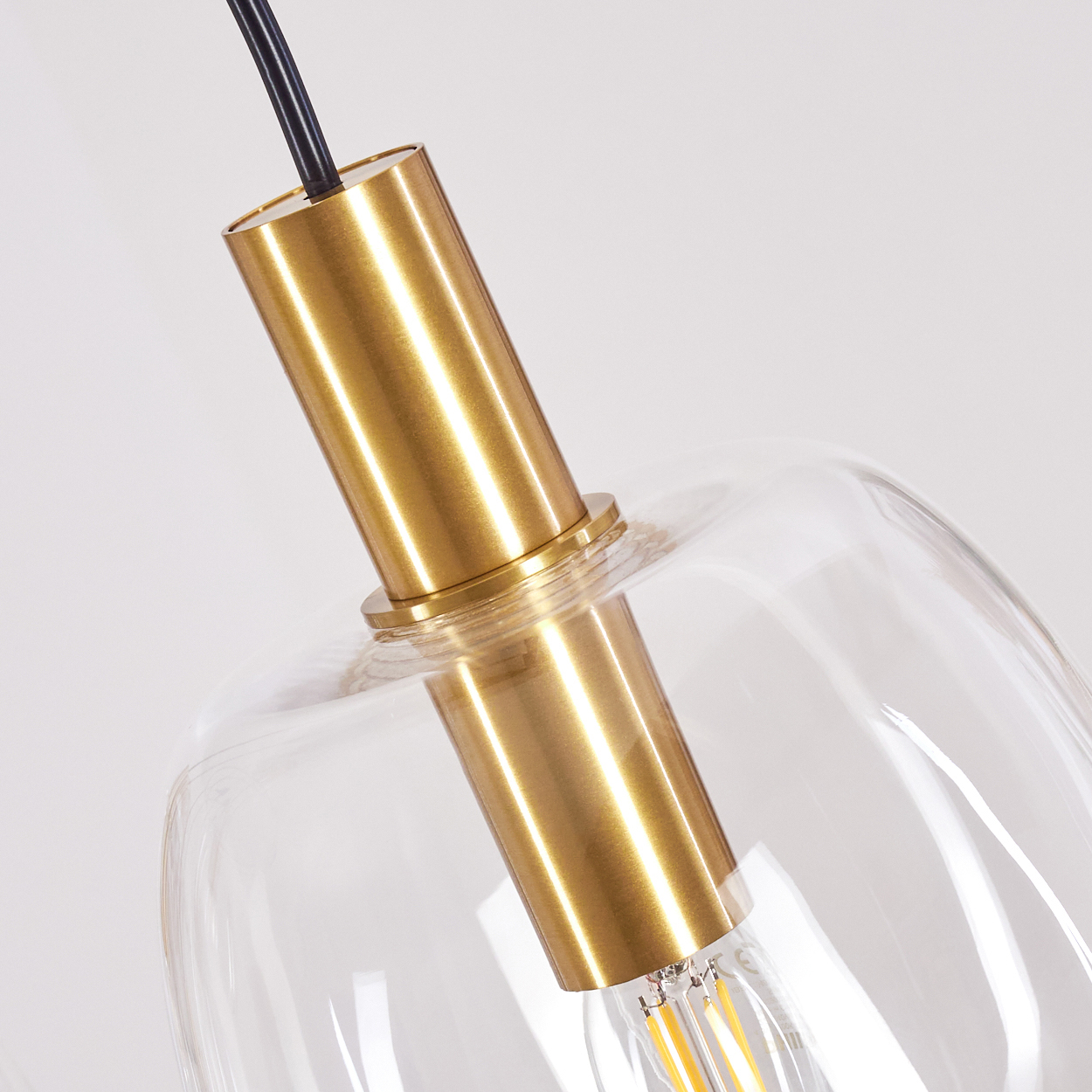 Nahaufnahme einer modernen Pendelleuchte. Sie hat einen klaren Glasschirm und einen goldfarbenen Metallrahmen. Ein schwarzes Kabel hängt von oben.