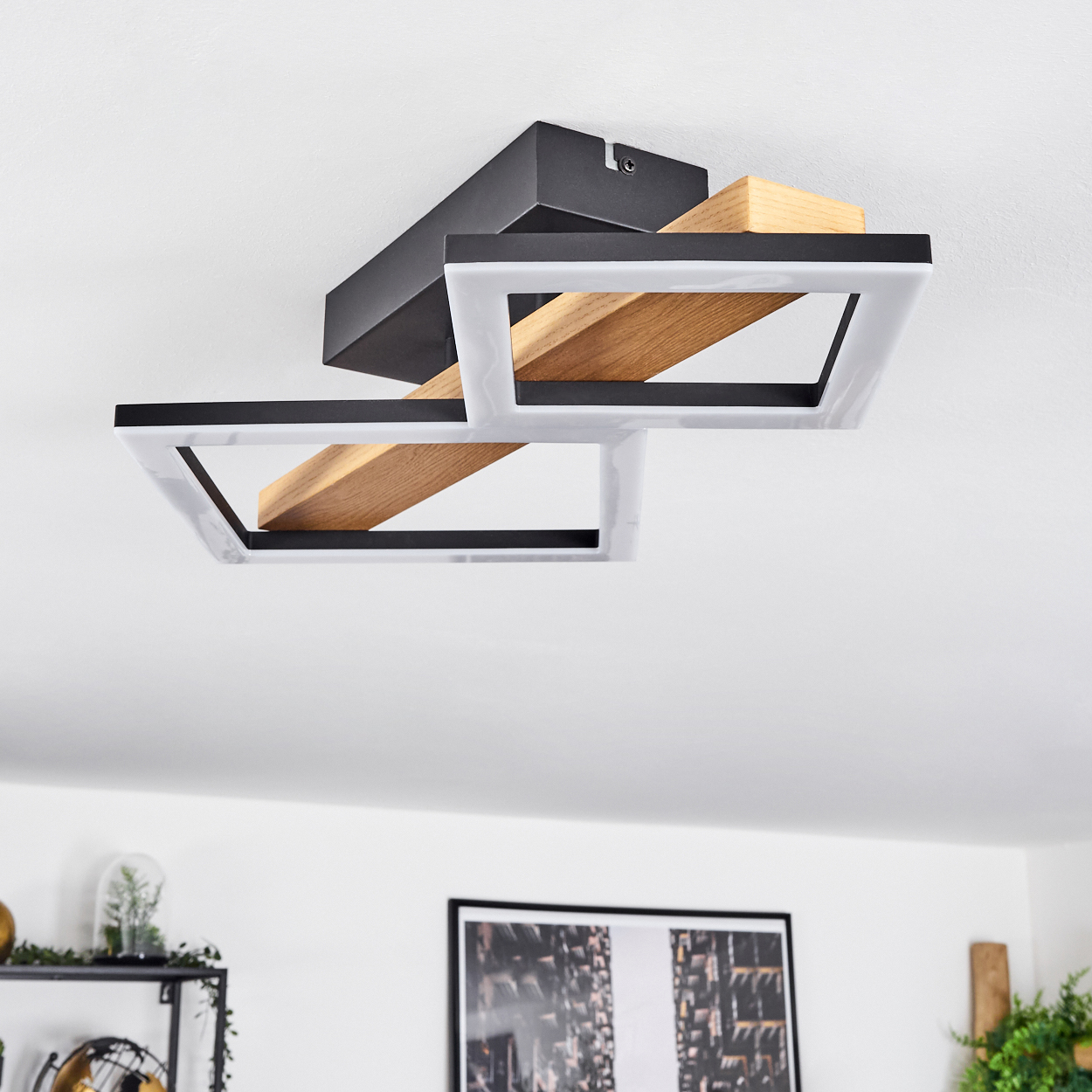 Moderne Deckenleuchte mit geometrischen Formen. Sie hat schwarze Rahmen, weißes Licht und Holzakzente. Sie ist in einem weißen Raum.