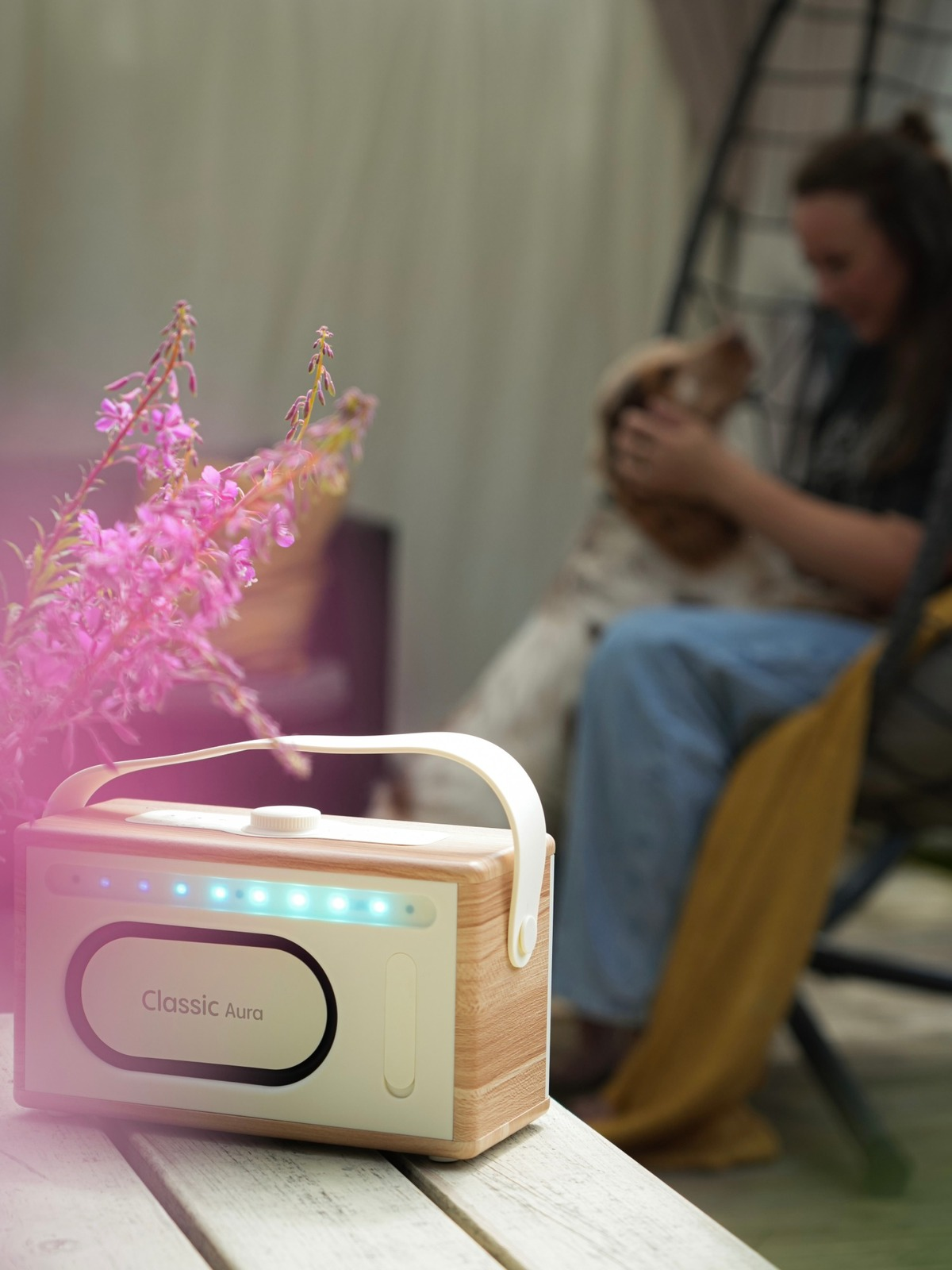 Une radio en bois avec le texte 'Classic Aura' est devant des fleurs roses. Une personne caresse un chien en arrière-plan.
