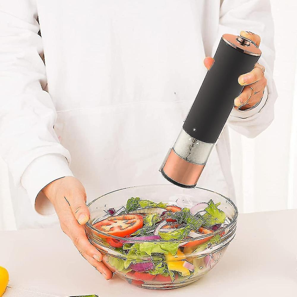 Eine Person hält einen schwarz-kupferfarbenen elektrischen Grinder über einer Glasschüssel mit Salat. Der Salat enthält Tomaten, Salat und Zwiebeln.