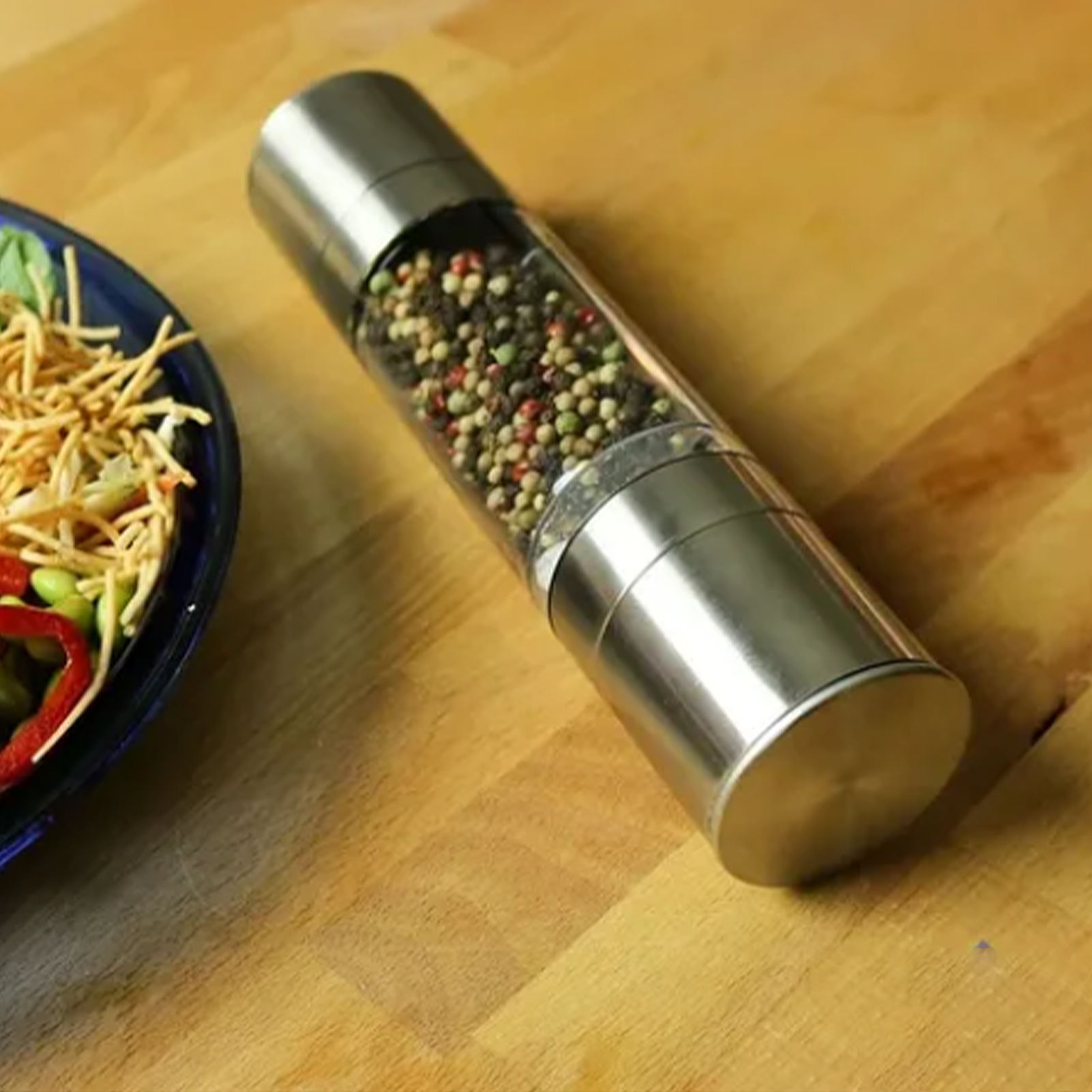 Eine Pfeffermühle auf einer Holzoberfläche, neben einer blauen Schüssel mit Salat. Die Mühle ist mit Pfefferkörnern gefüllt.