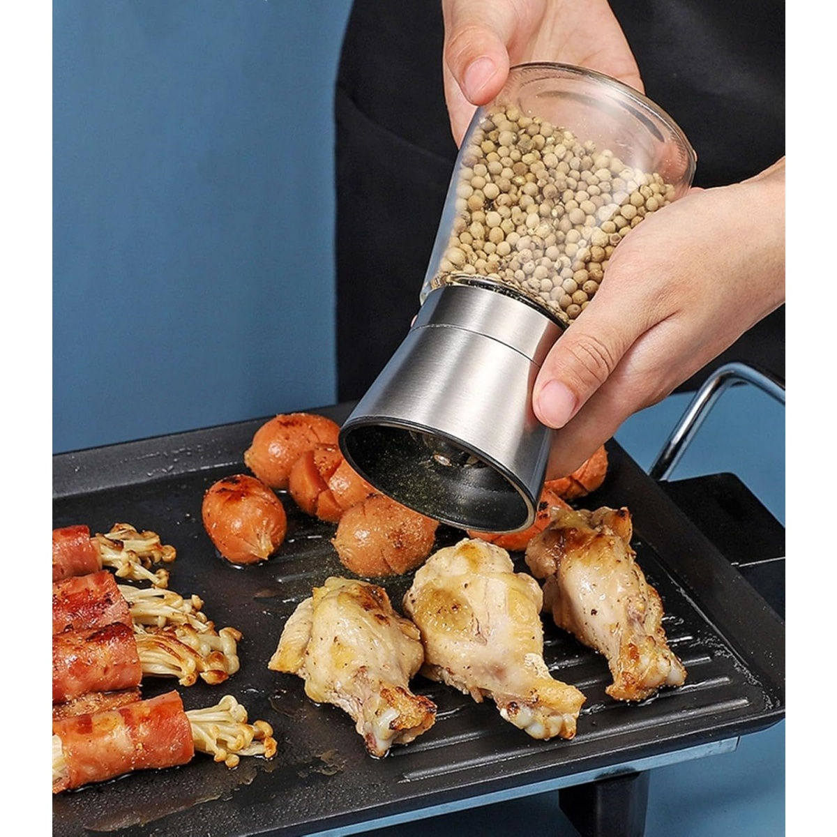 Hände mahlen Pfeffer auf gegrillte Hähnchenflügel und Würstchen. Speck-umwickelte Enoki-Pilze sind dabei.