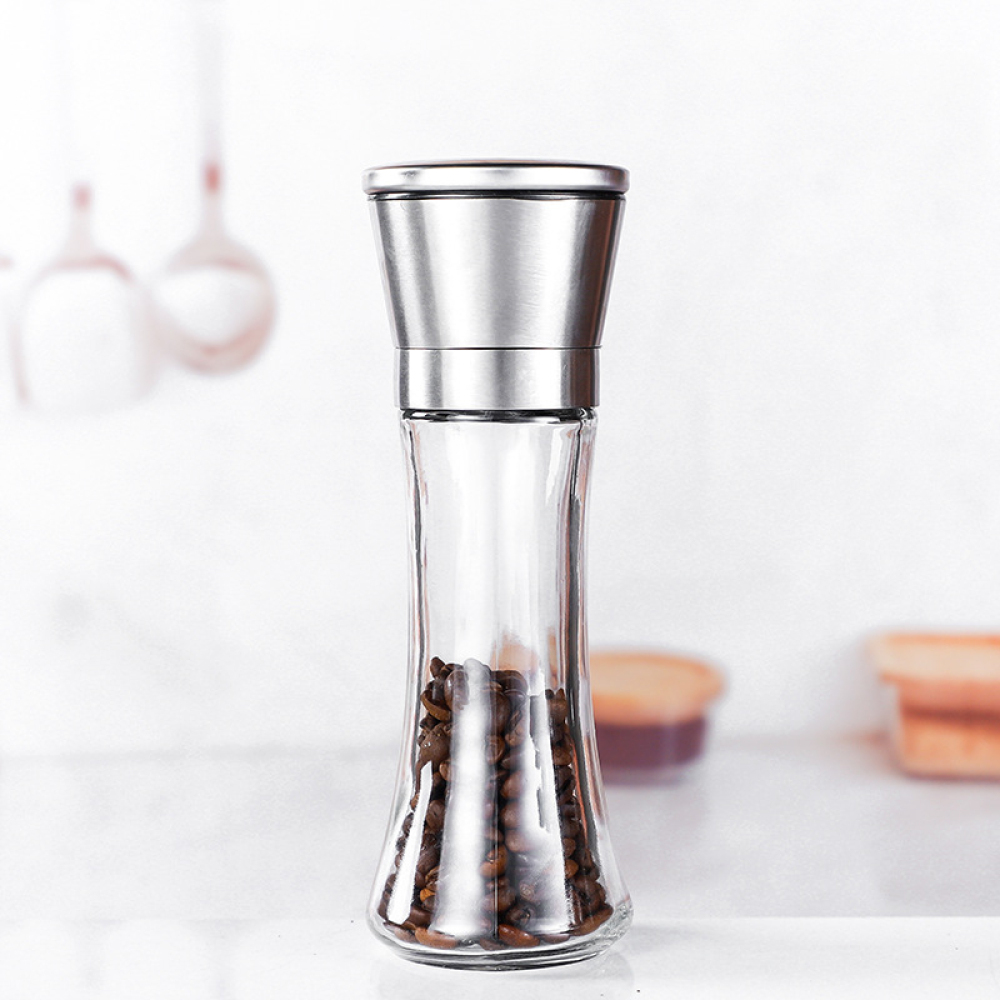 Eine klare Pfeffermühle aus Glas mit Edelstahlaufsatz. Sie enthält Kaffeebohnen und steht auf einer weißen Oberfläche.