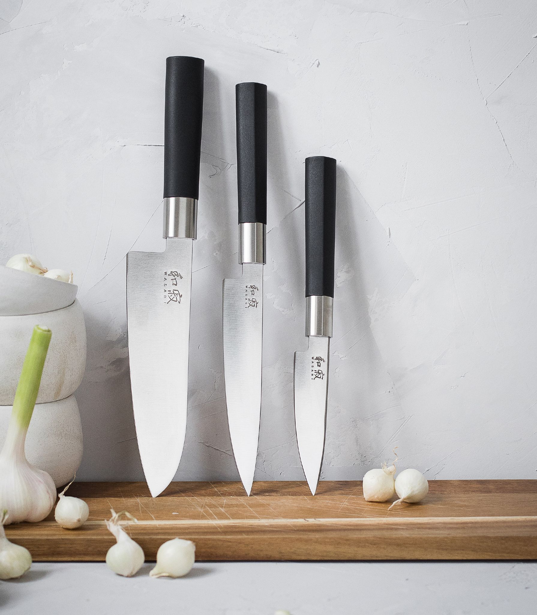 Drei Messer mit schwarzen Griffen stehen vor einer grauen Wand. Knoblauch und Frühlingszwiebeln liegen auf einem Holzschneidebrett.