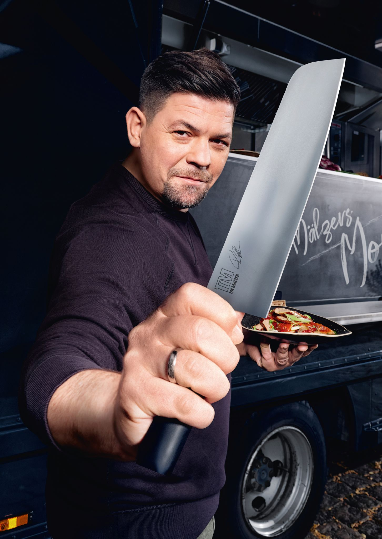 Ein Mann hält ein großes Messer und einen Teller mit Essen. Er steht vor einem Foodtruck mit dem Namen Dälzers Mo.