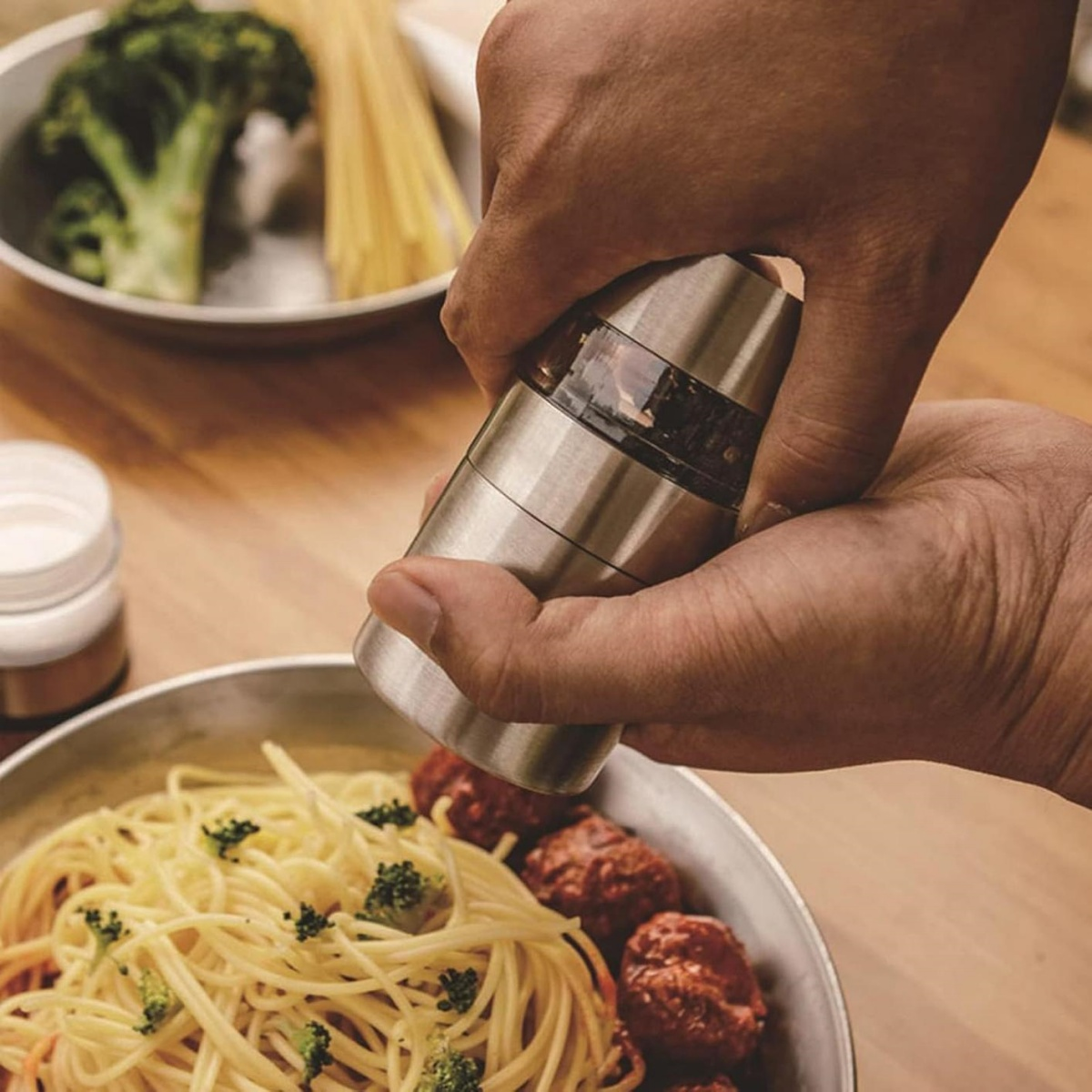 Eine Person mahlt Pfeffer auf Spaghetti mit Fleischbällchen. Brokkoli und Nudeln sind im Hintergrund.