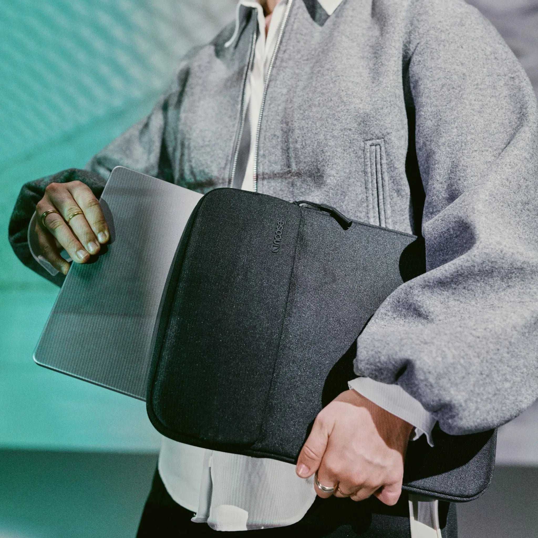 Persona che tiene in mano un laptop e una custodia nera. Indossa una giacca grigia e una camicia bianca.