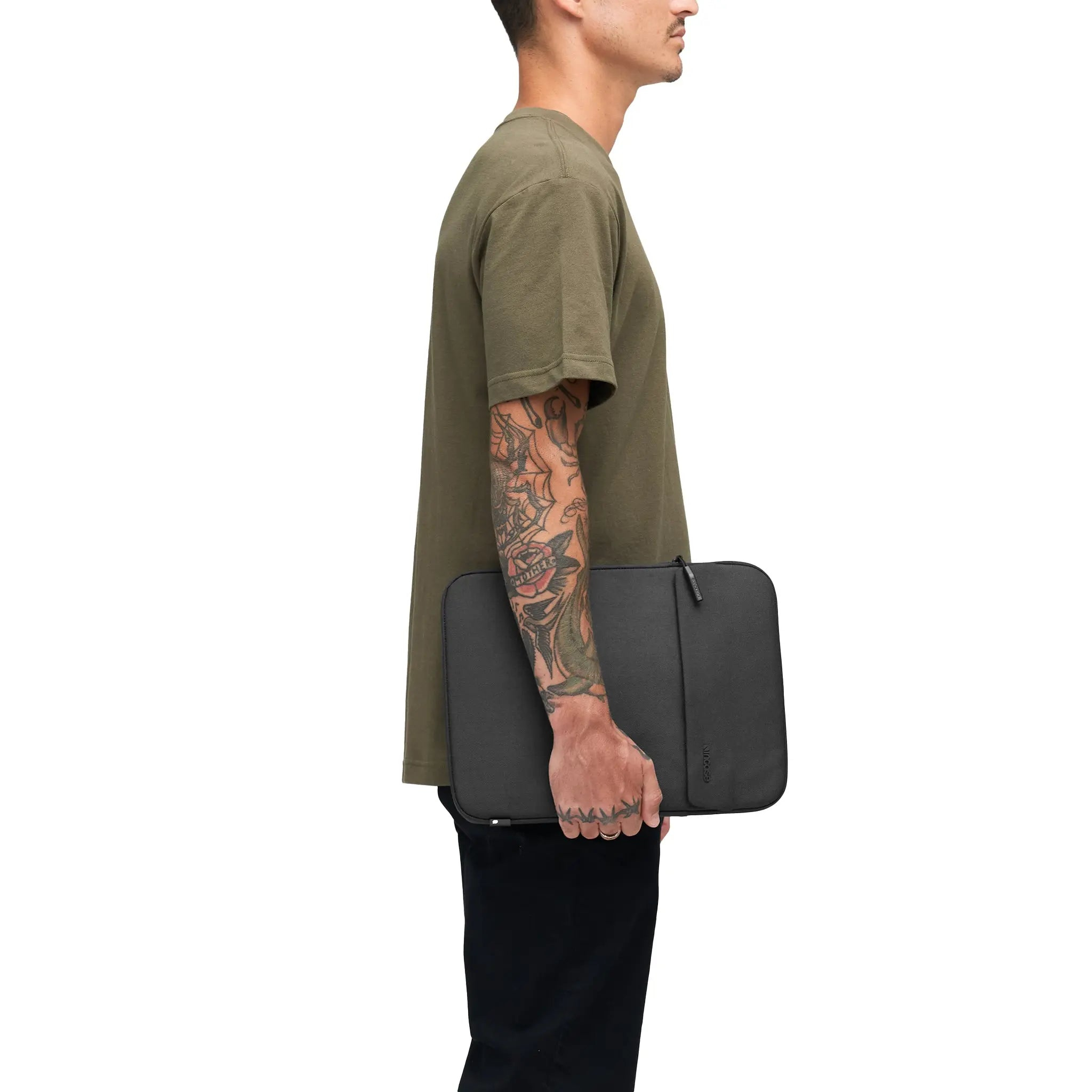 Un uomo con camicia verde, pantaloni neri, tiene una custodia nera per laptop, tatuaggi sul braccio.