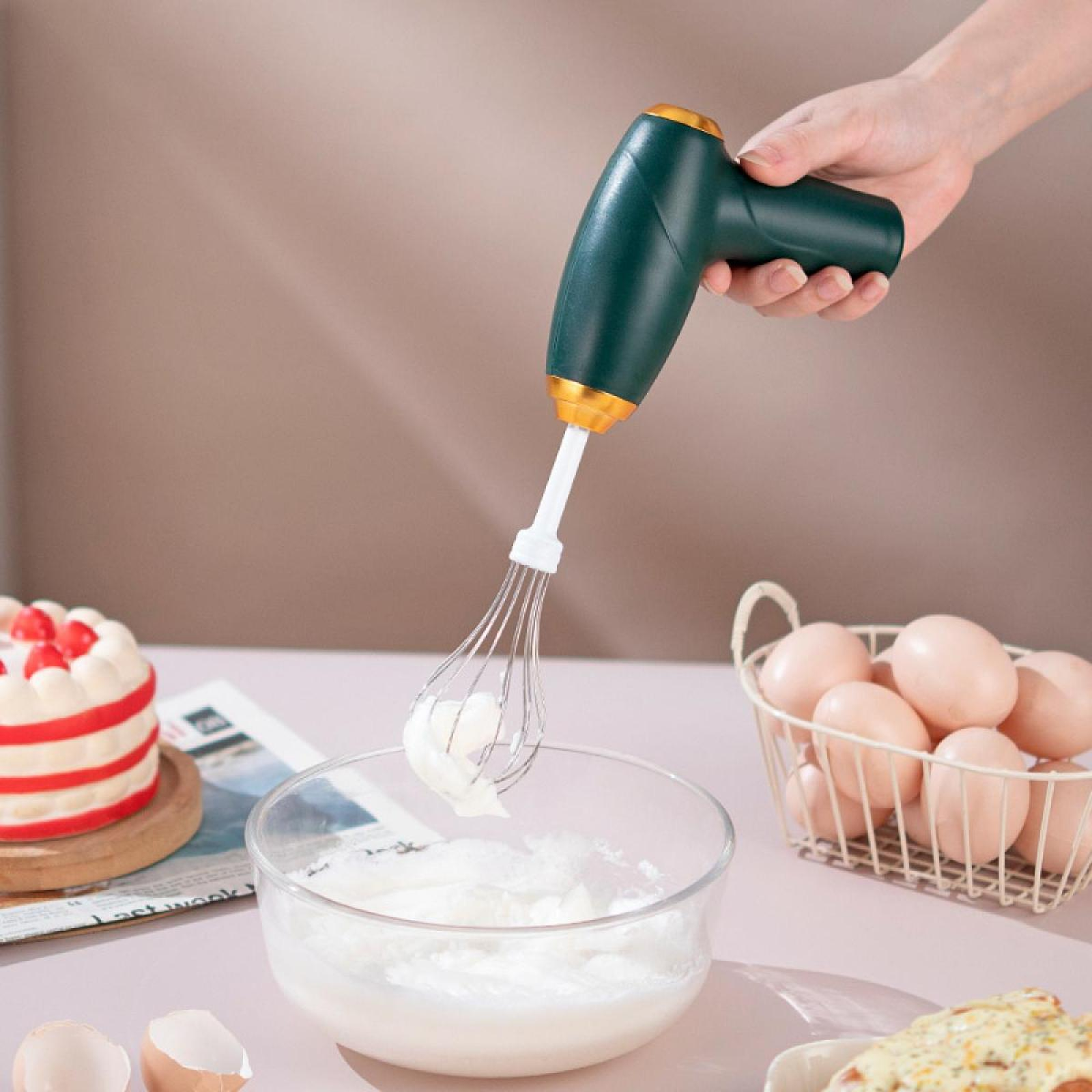 Hand hält einen grünen und goldenen Mixer, der Eiweiß in einer Glasschüssel aufschlägt. Eier, Kuchen und Essen stehen auf einem Tisch.