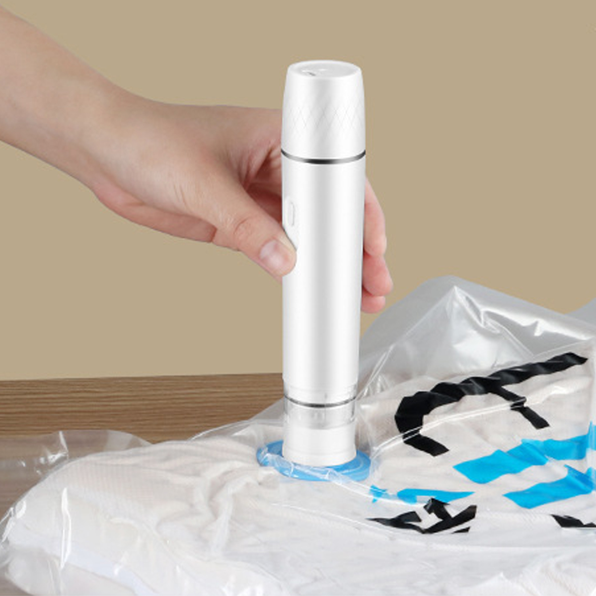 Eine Hand hält einen weißen Vakuumversiegler über einer Tasche mit Kleidung. Der Beutel ist transparent, und der Hintergrund ist eine beige Wand.