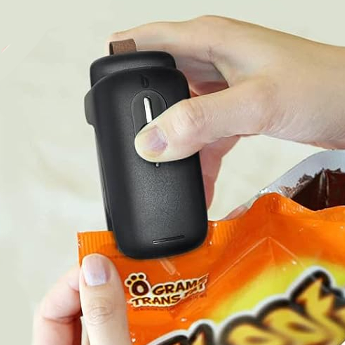 Eine Person verschließt einen orangenen Snackbeutel mit einem schwarzen Handversiegeler. Der Beutel hat Text und einen Pfotenabdruck. Die Hand hält den Beutel.