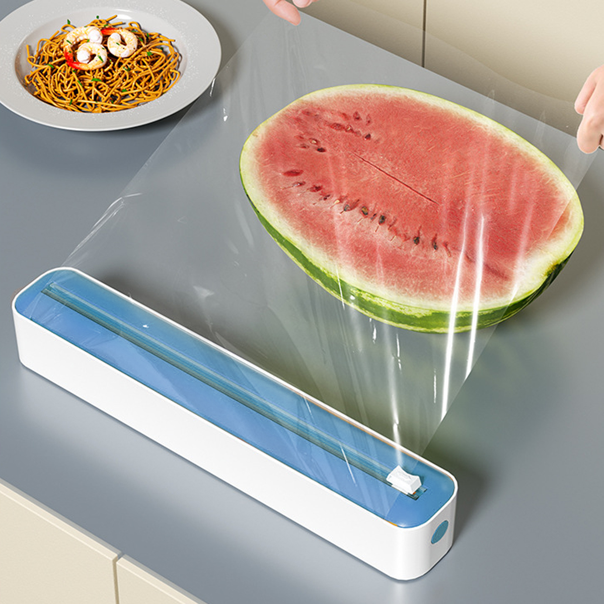 Eine halbe Wassermelone wird in Frischhaltefolie eingewickelt. Spaghetti mit Garnelen liegen auf einem Teller. Ein Frischhaltefolienspender steht auf dem Tisch.