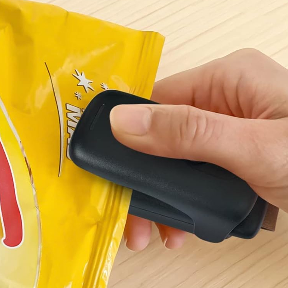 Eine Hand verschließt einen gelben Chip-Beutel mit einem schwarzen Versiegeler. Der Beutel hat weißen Text. Der Hintergrund ist eine Holzoberfläche.