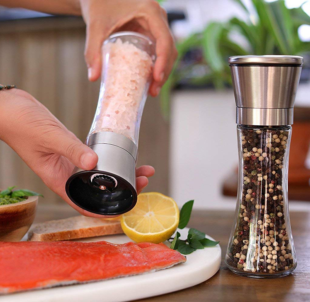 Eine Person hält eine Salzmühle neben einer Pfeffermühle. Lachs, Zitrone und Kräuter liegen auf einem Schneidebrett.
