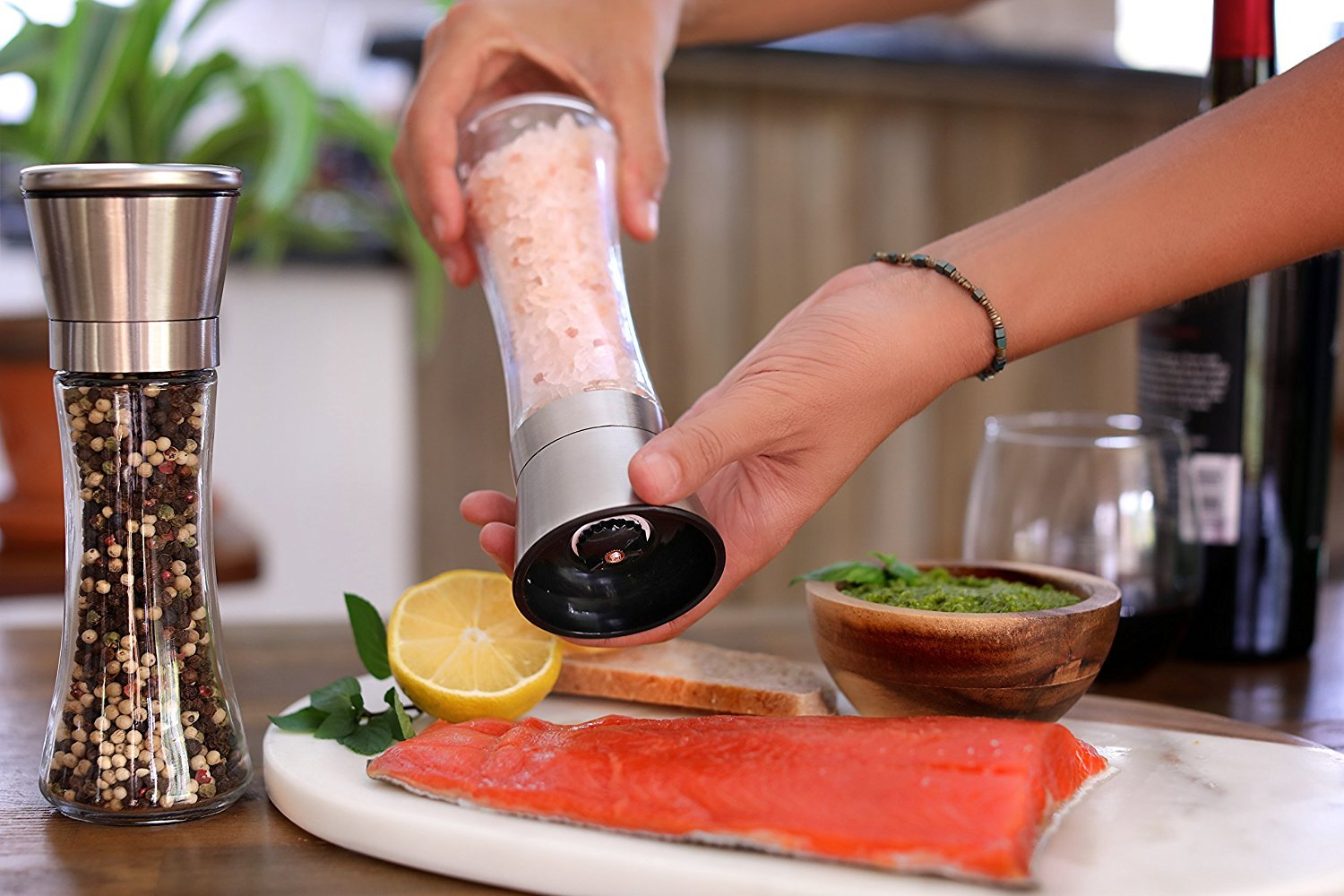 Hände mahlen Salz über einem Teller mit Lachs, Zitrone, Pesto und Brot. Pfeffermühle links, Weinflasche und Glas im Hintergrund.
