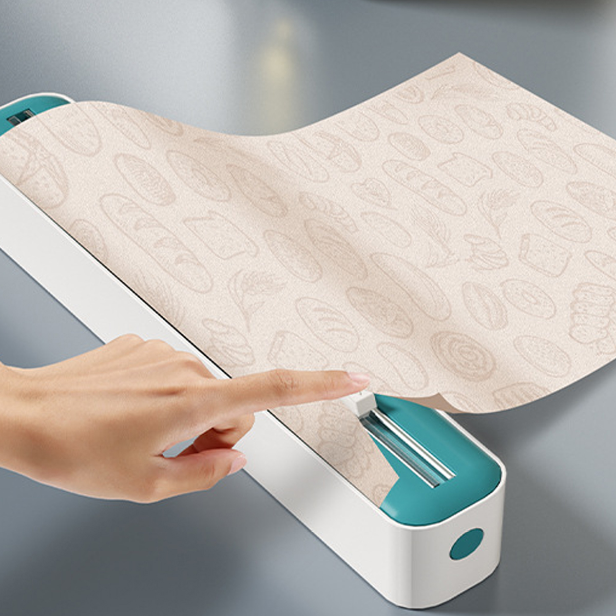 Eine Hand drückt einen Schneider auf einer Papierrolle mit Brotmustern. Das Gerät ist weiß und türkis. Der Hintergrund ist grau.