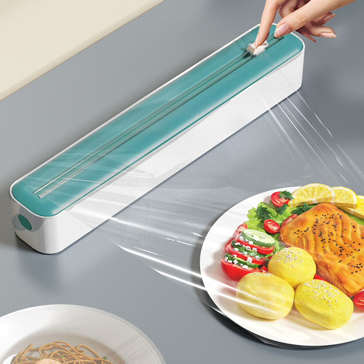 Eine Hand, die Plastikfolie über einen Teller mit Essen, darunter Lachs, Tomaten und Zitronen, aus einem teal-weißen Spender abgibt.