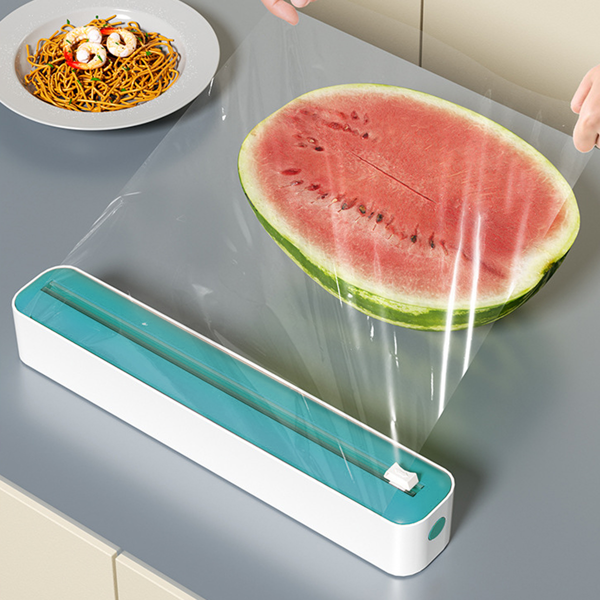 Eine Wassermelonen-Hälfte wird in Frischhaltefolie eingewickelt. Spaghetti mit Garnelen auf einem Teller. Frischhaltefolien-Spender auf dem Tisch.