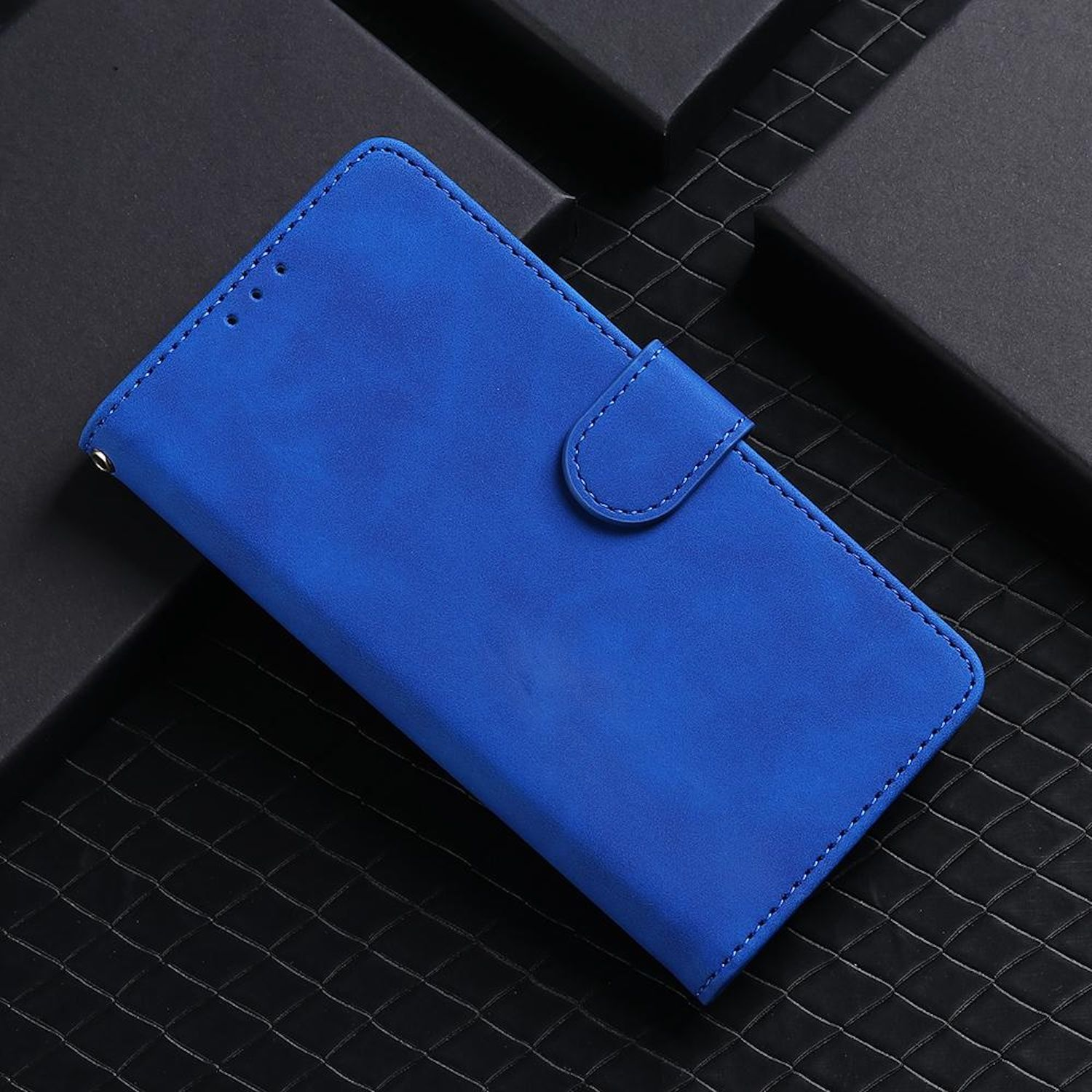 Eine blaue Handyhülle mit Magnetverschluss auf einer strukturierten schwarzen Oberfläche neben einer schwarzen Box. Die Hülle ist offen.