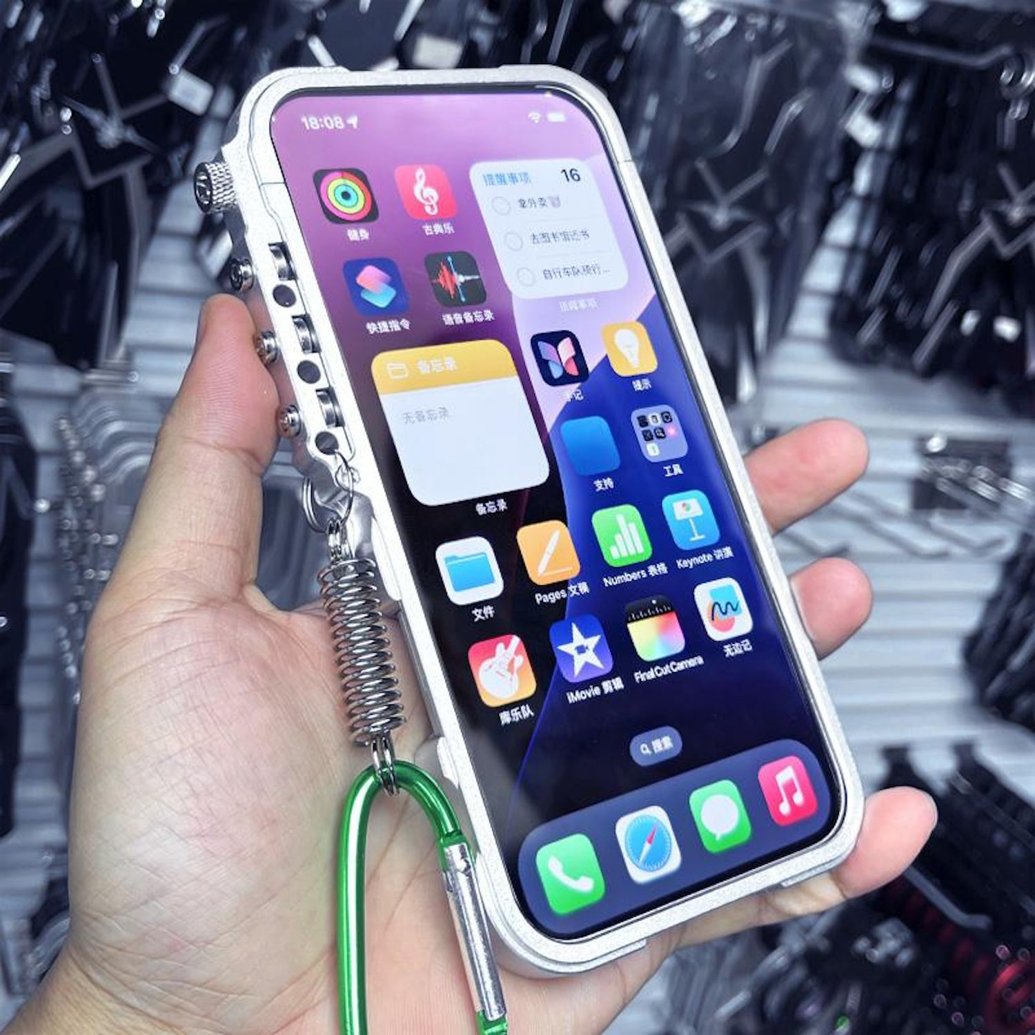 Una mano sostiene un teléfono en una funda plateada. La pantalla muestra iconos de aplicaciones sobre un fondo morado. Un resorte y clip verdes adjuntos.