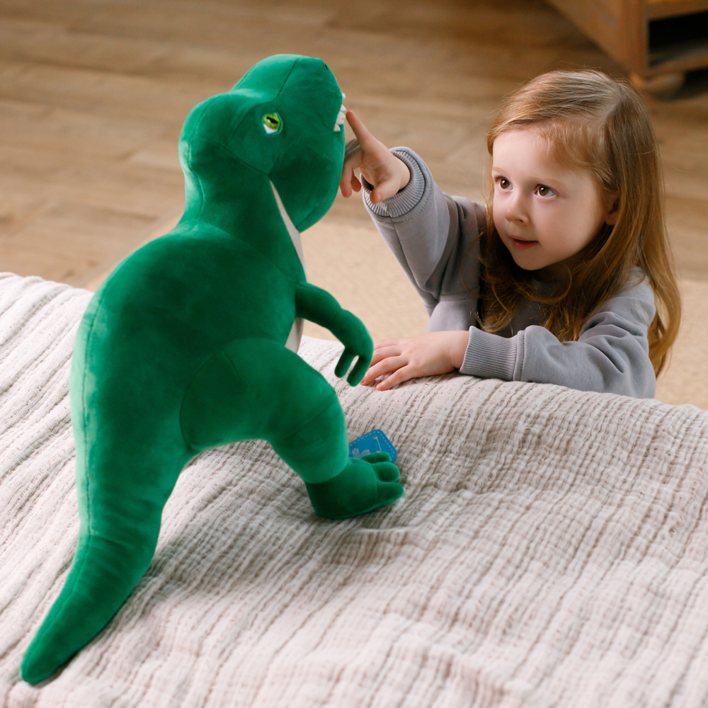 Ein junges Mädchen berührt ein grünes Dinosaurierspielzeug. Das Spielzeug liegt auf einem Bett mit einer weißen Decke. Das Mädchen trägt einen grauen Pullover.