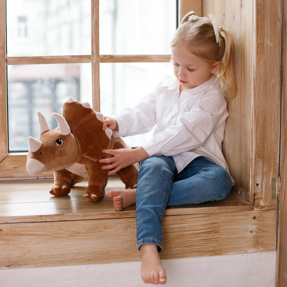 Ein junges Mädchen in Jeans und einem weißen Hemd sitzt an einem Fenster und berührt ein braunes Triceratops-Plüschtier.