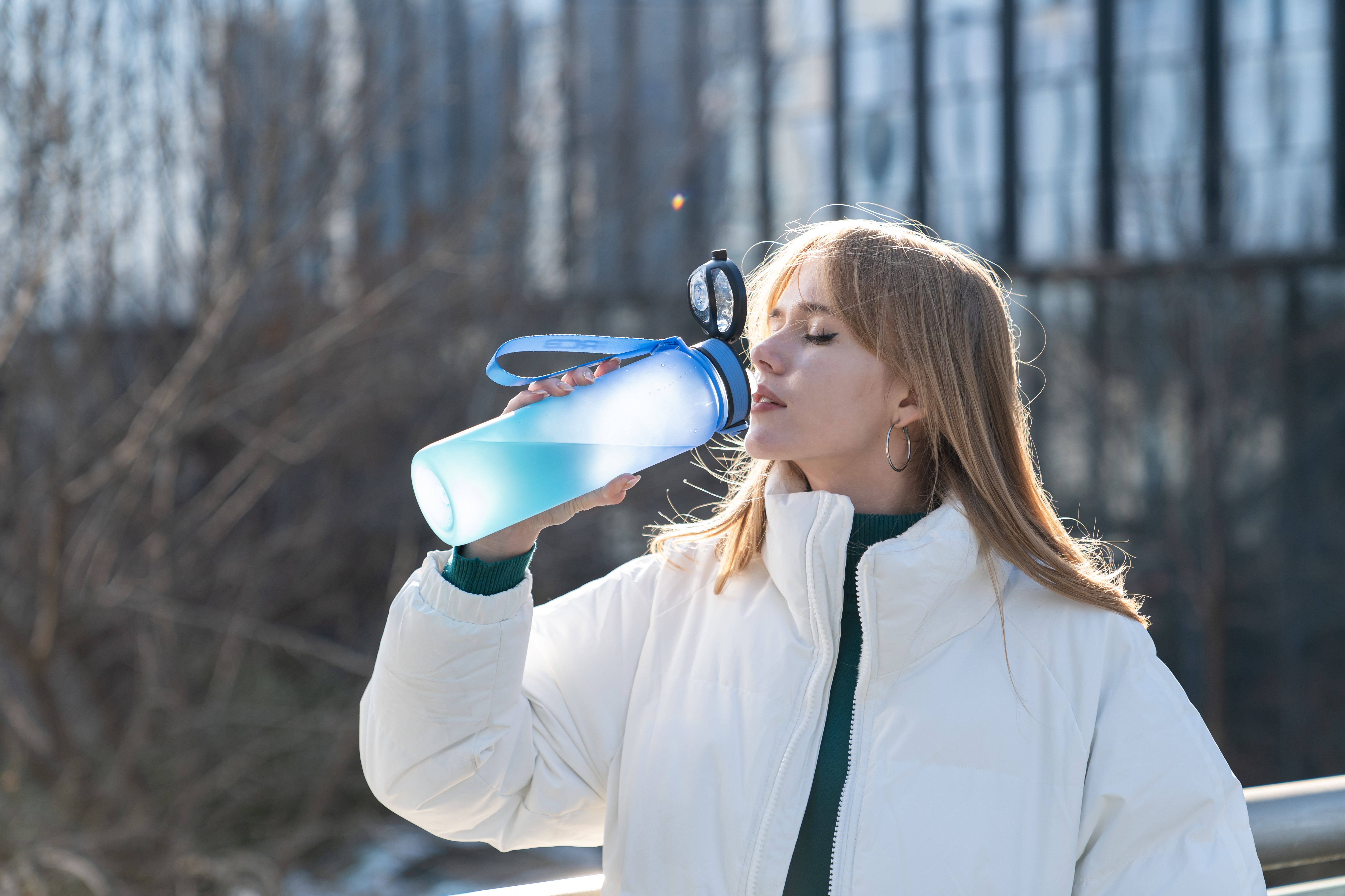Een vrouw drinkt uit een blauwe waterfles. Ze draagt een witte jas en staat buiten met gebouwen op de achtergrond.