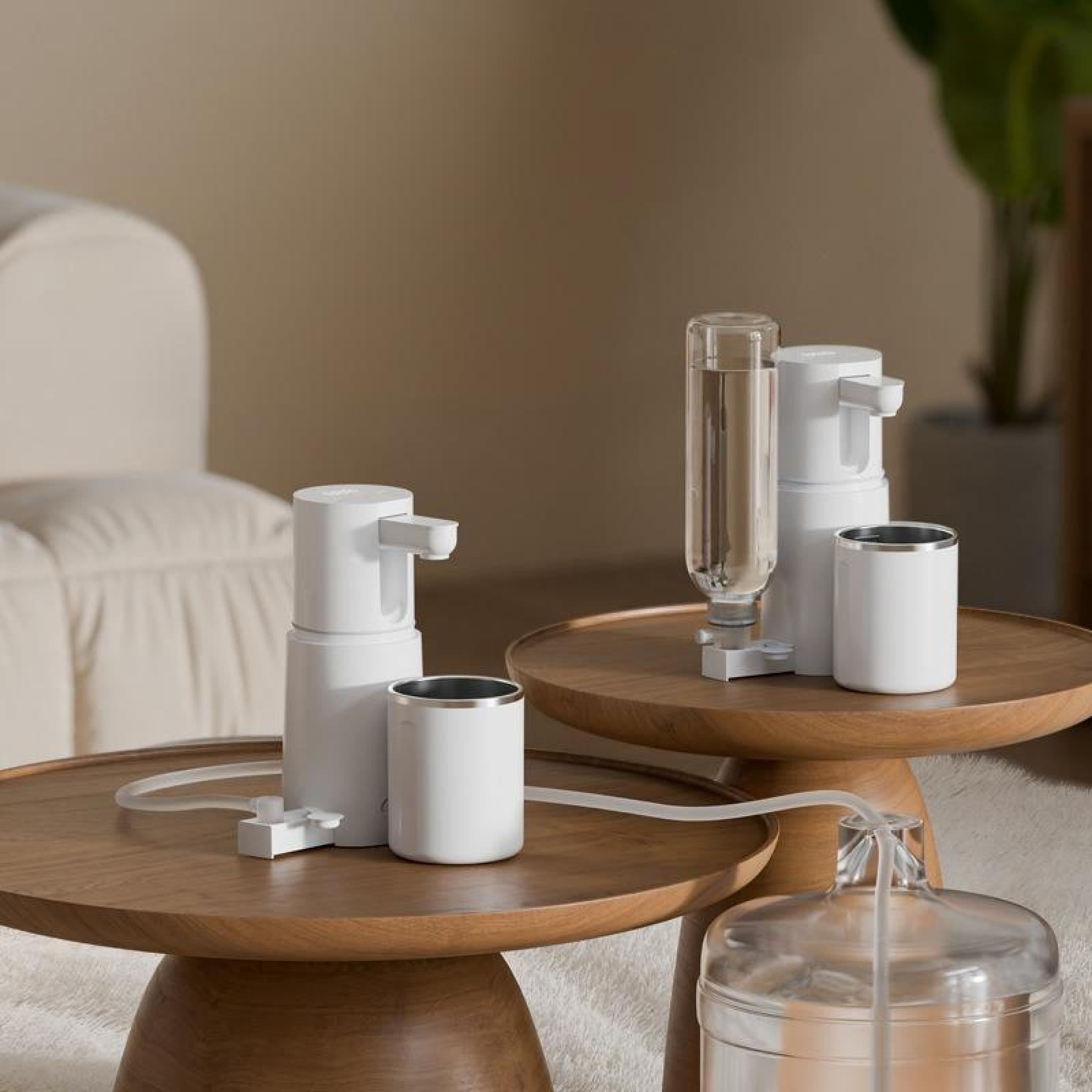 Weiße Wasserspender auf Holztischen. Klare Wasserflasche und Becher. Beige Sofa und Teppich im Hintergrund.