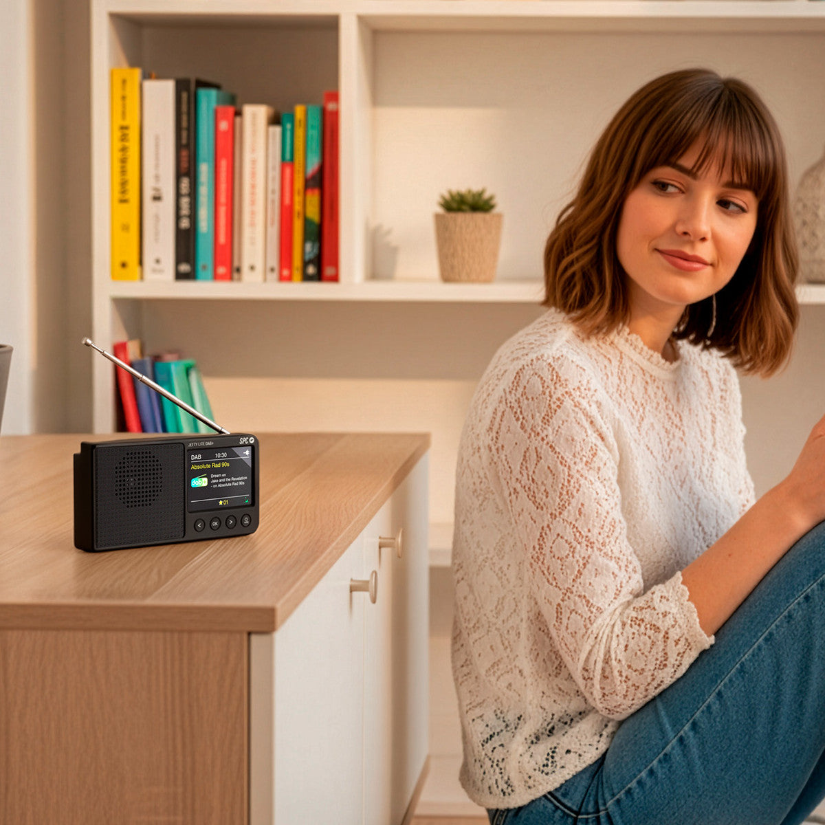 Eine Frau sitzt in der Nähe eines Radios auf einem Tisch. Bücher und eine Pflanze sind im Hintergrund. Sie trägt ein weißes Spitzenshirt.