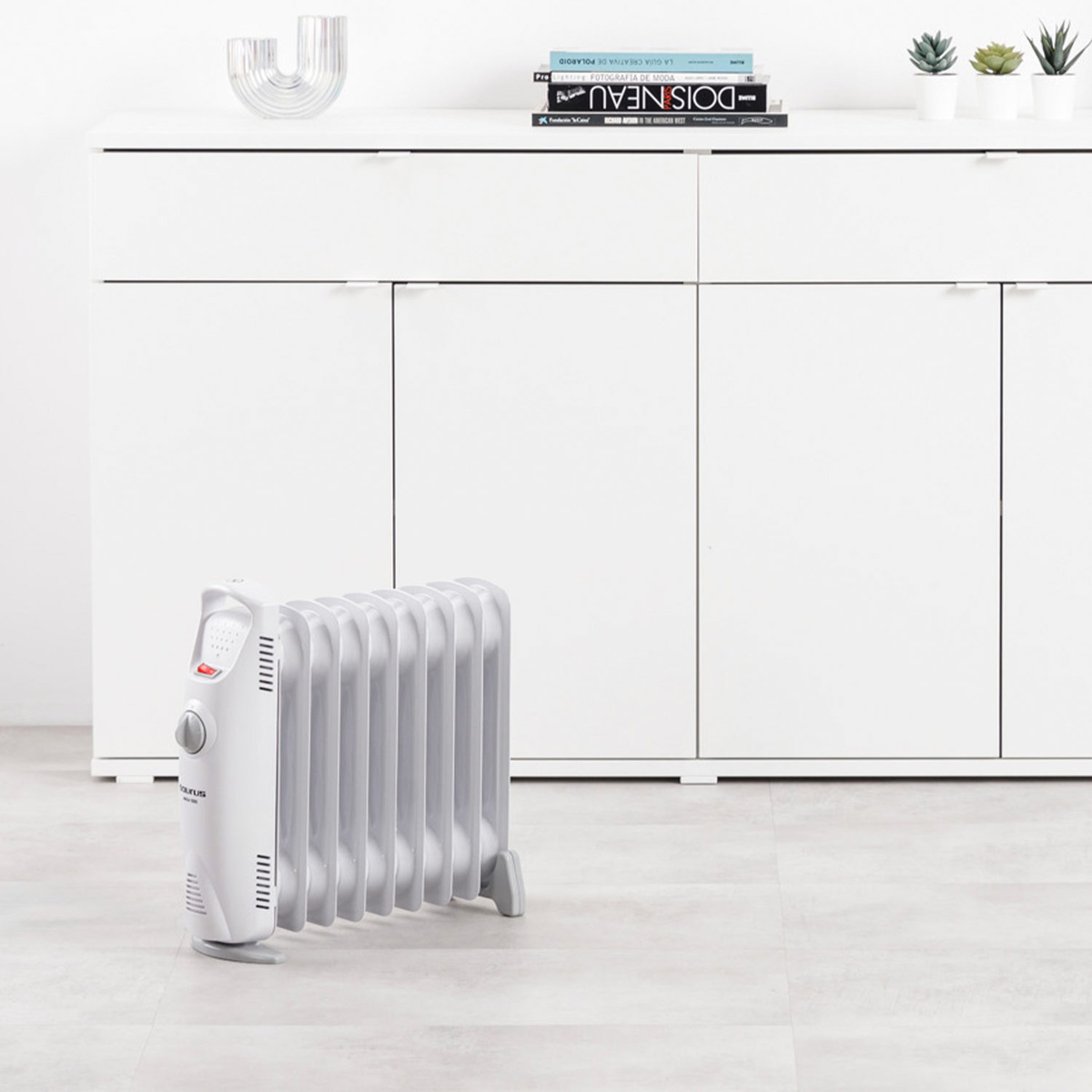 Biało-szary grzejnik olejowy stoi przed białą szafką. Na górze szafki są książki i rośliny.