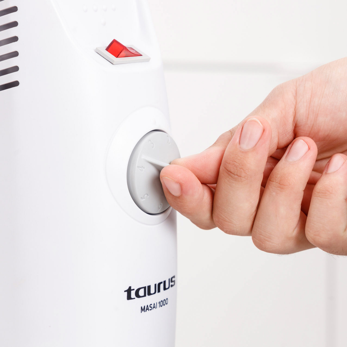 Ręka reguluje szare pokrętło na białym grzejniku. Widoczny jest czerwony przełącznik. Widnieje nazwa marki 'taurus'.