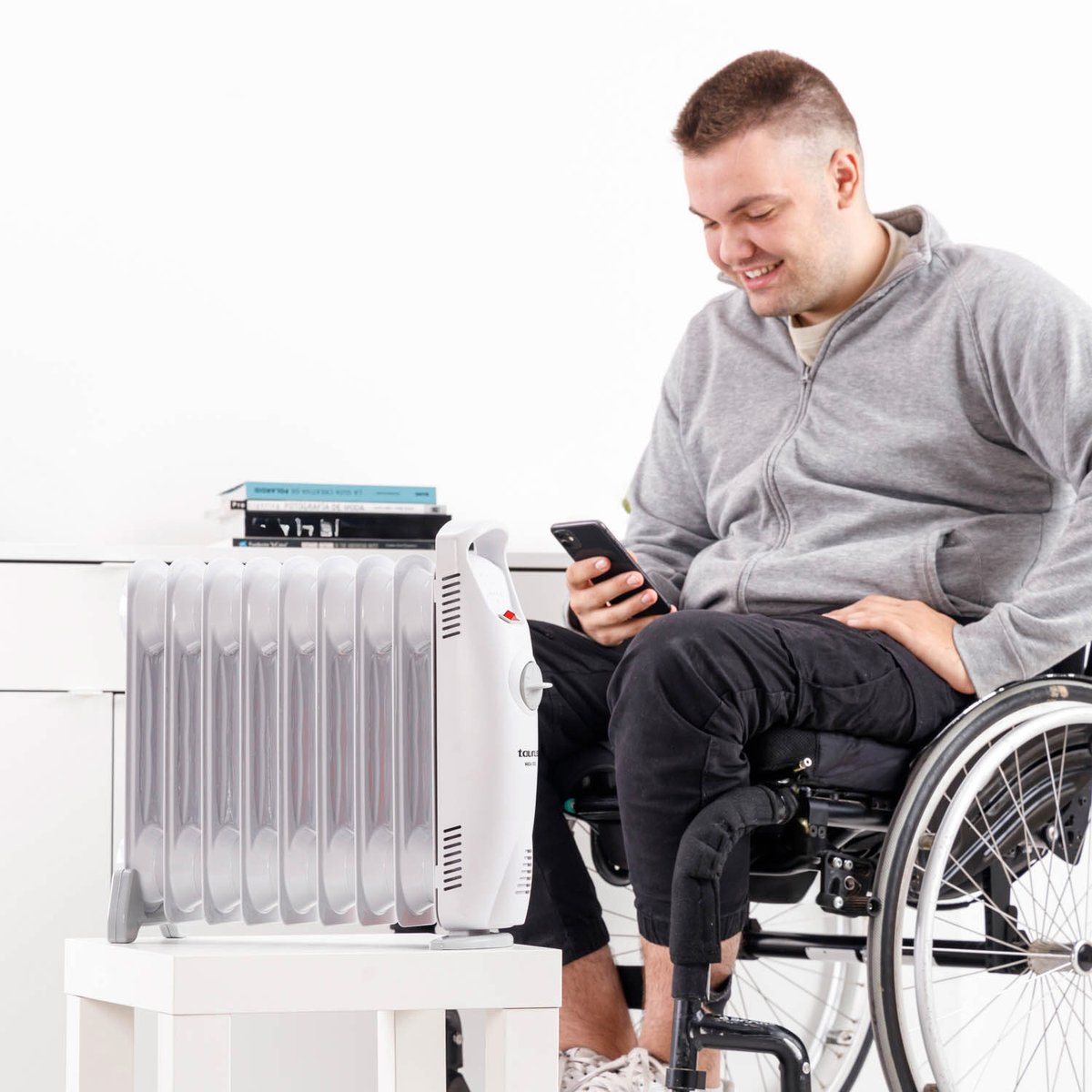 Mężczyzna na wózku inwalidzkim uśmiecha się, patrząc na telefon. Biały grzejnik stoi na białym stole. Książki.