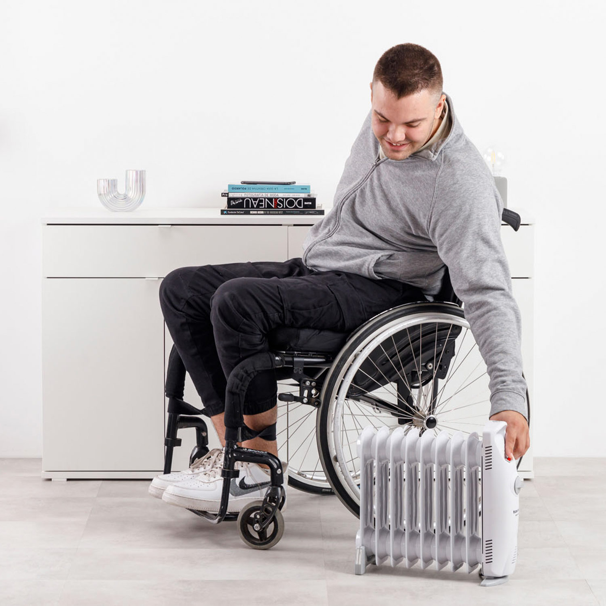 Mężczyzna na wózku inwalidzkim sięga po biały grzejnik na podłodze. Ma na sobie szarą kurtkę i czarne spodnie. Książki na szafce.