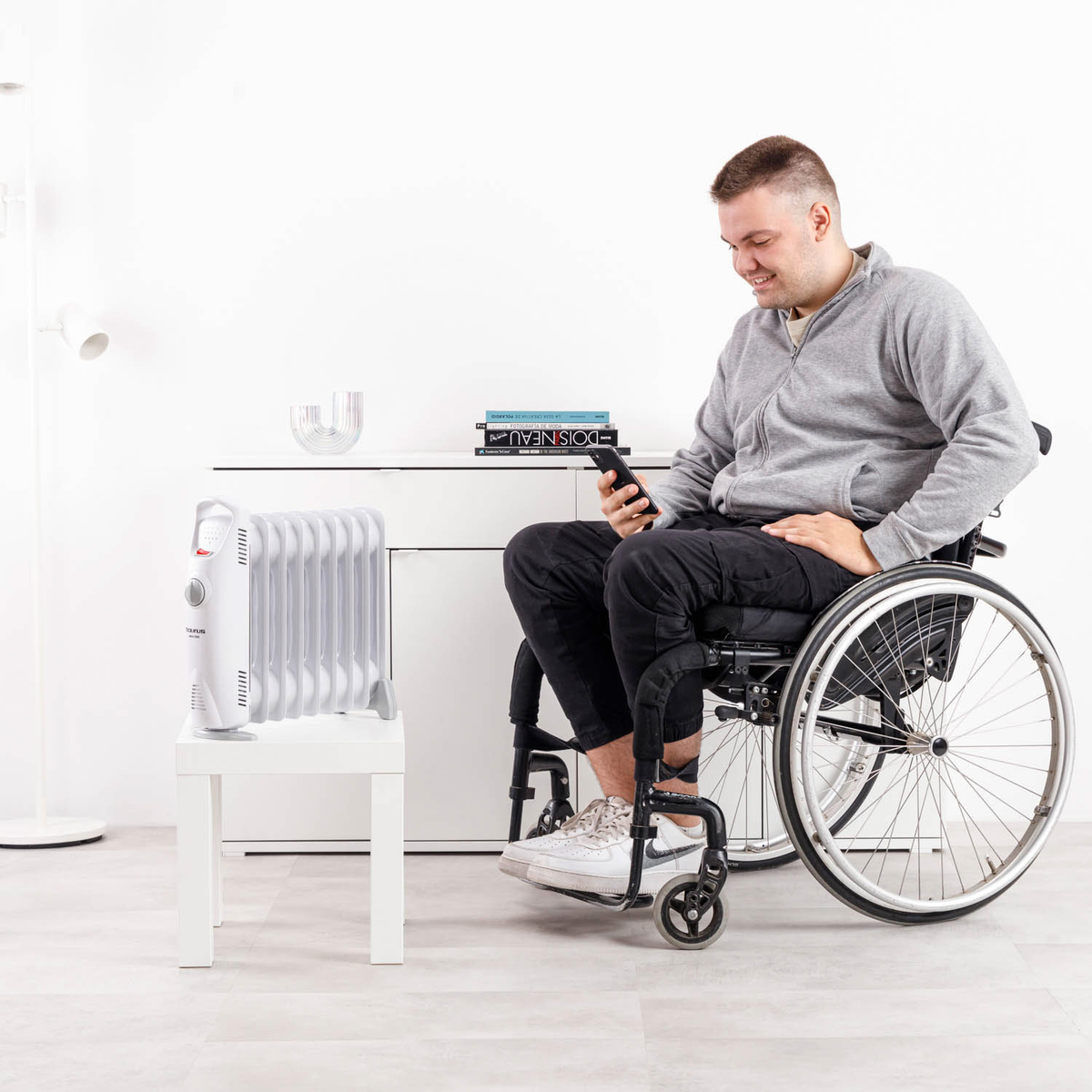 Mężczyzna na wózku inwalidzkim uśmiecha się patrząc na telefon. Biały grzejnik na małym stoliku. Biała szafka i książki w tle.