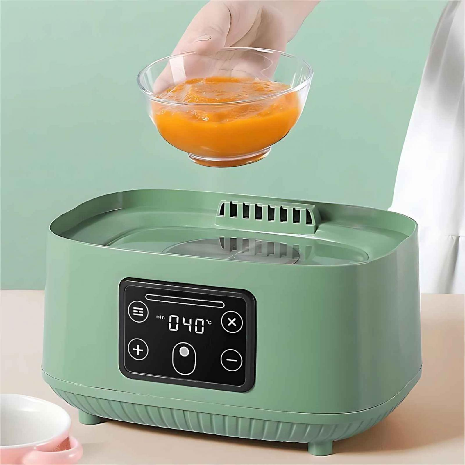 Eine Hand hält eine Glasschale mit orangefarbenem Babynahrung über einem grünen Essenswärmer.