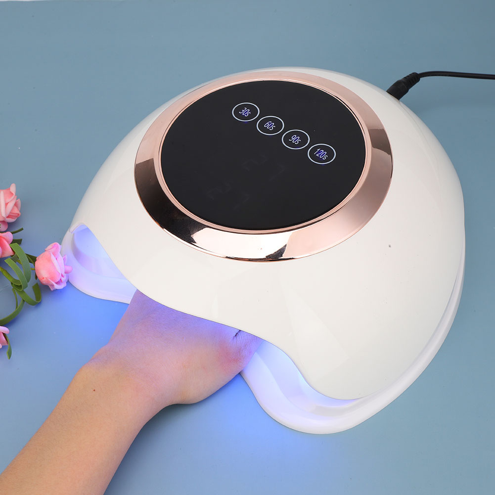 Eine weiße Nagellampe mit einer Hand darin. Die Lampe hat eine Digitalanzeige und roségoldfarbene Akzente. Links sind rosa Blumen.