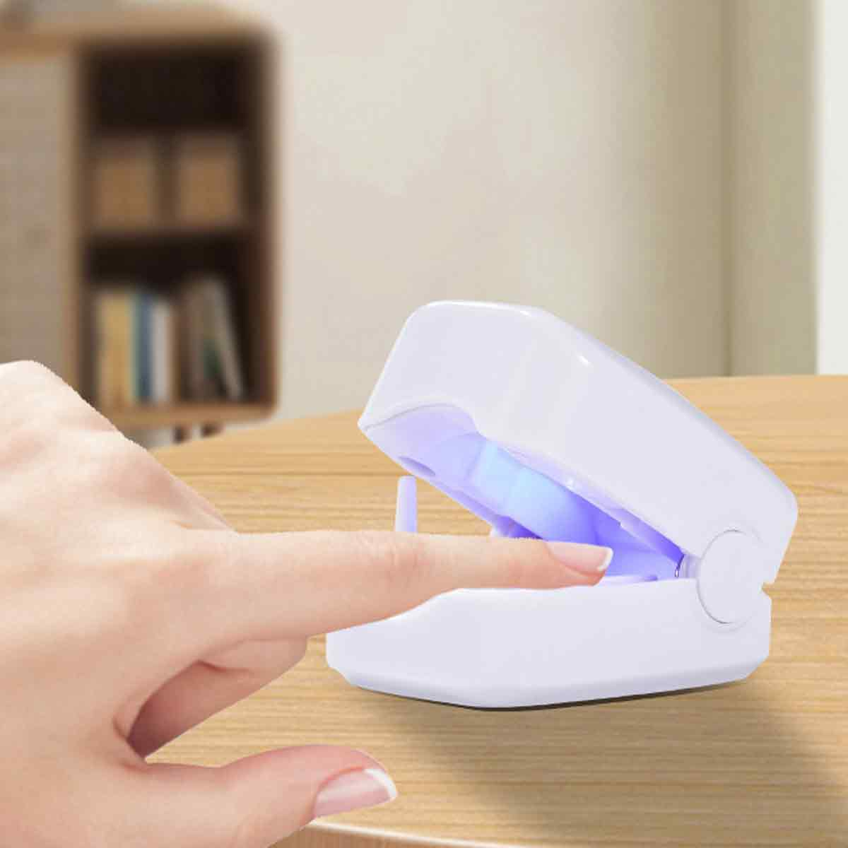 Eine Hand legt einen Finger in eine weiße Nagellampe. Die Lampe ist geöffnet und strahlt blaues Licht aus. Ein Holztisch und unscharfer Hintergrund.