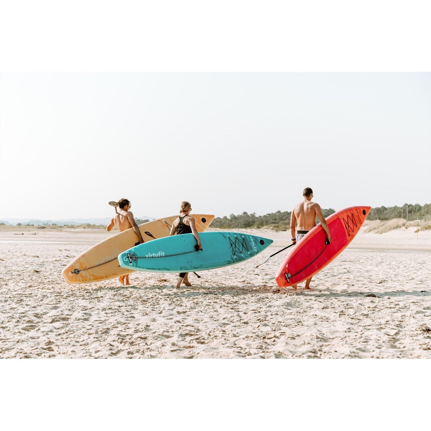 Drei Personen tragen Surfbretter an einem Sandstrand. Surfbretter sind gelb, türkis und rot. Der Himmel ist klar.