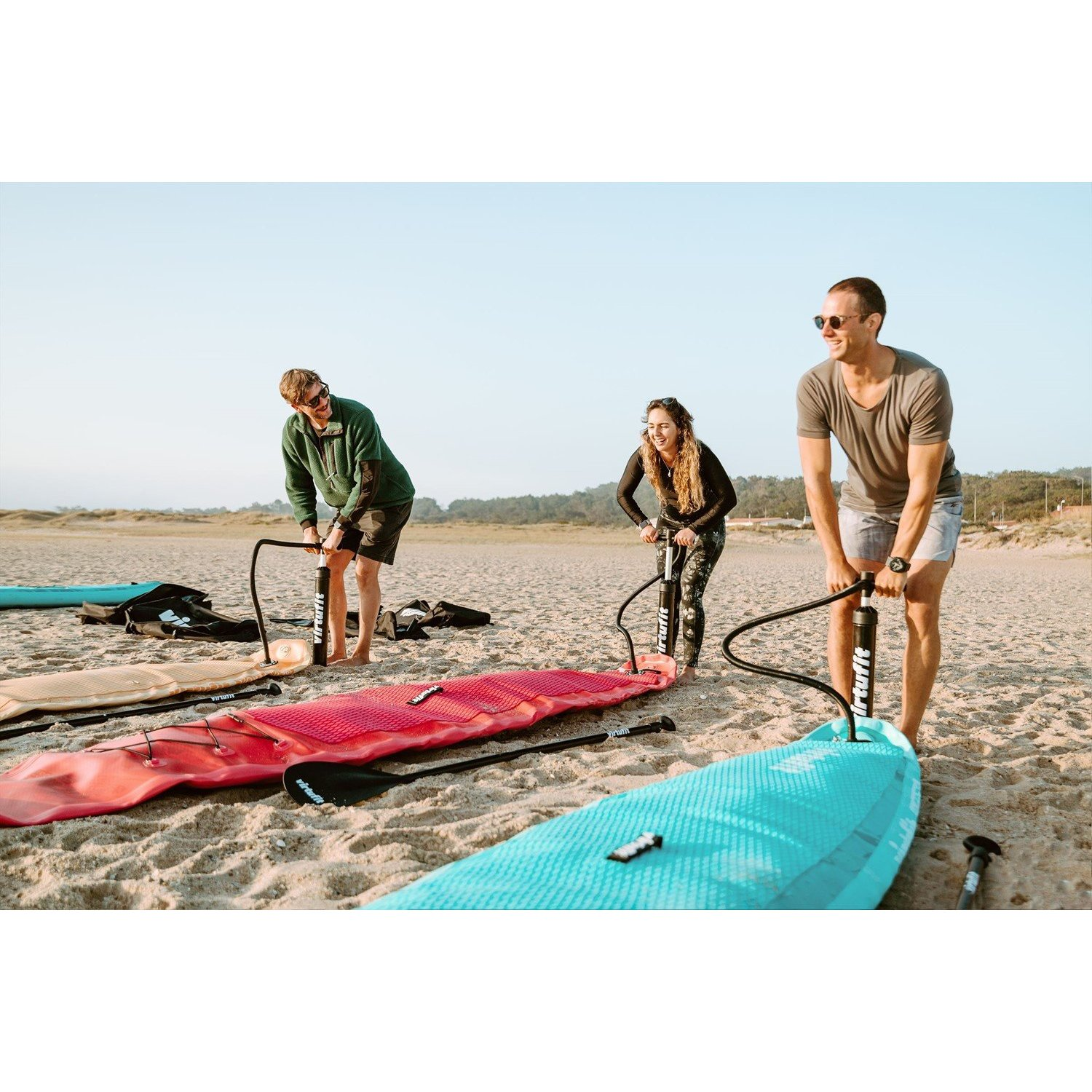 Drei Personen pumpen auf einem Sandstrand Paddleboards auf. Einer in Grün, einer in Schwarz und einer in Grau. Blauer Himmel und Bäume im Hintergrund.