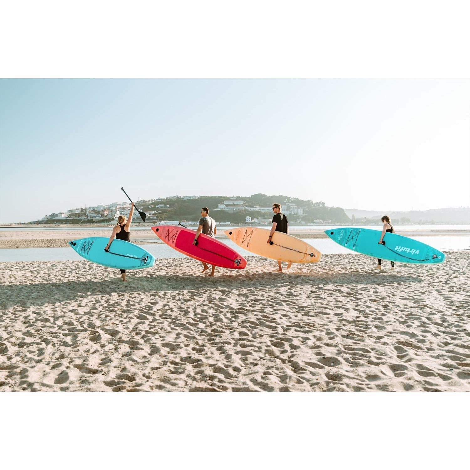 Vier Personen tragen bunte Surfbretter an einem Sandstrand. Eine Frau hält ein Paddel. Im Hintergrund sind eine Stadt und klarer Himmel zu sehen.