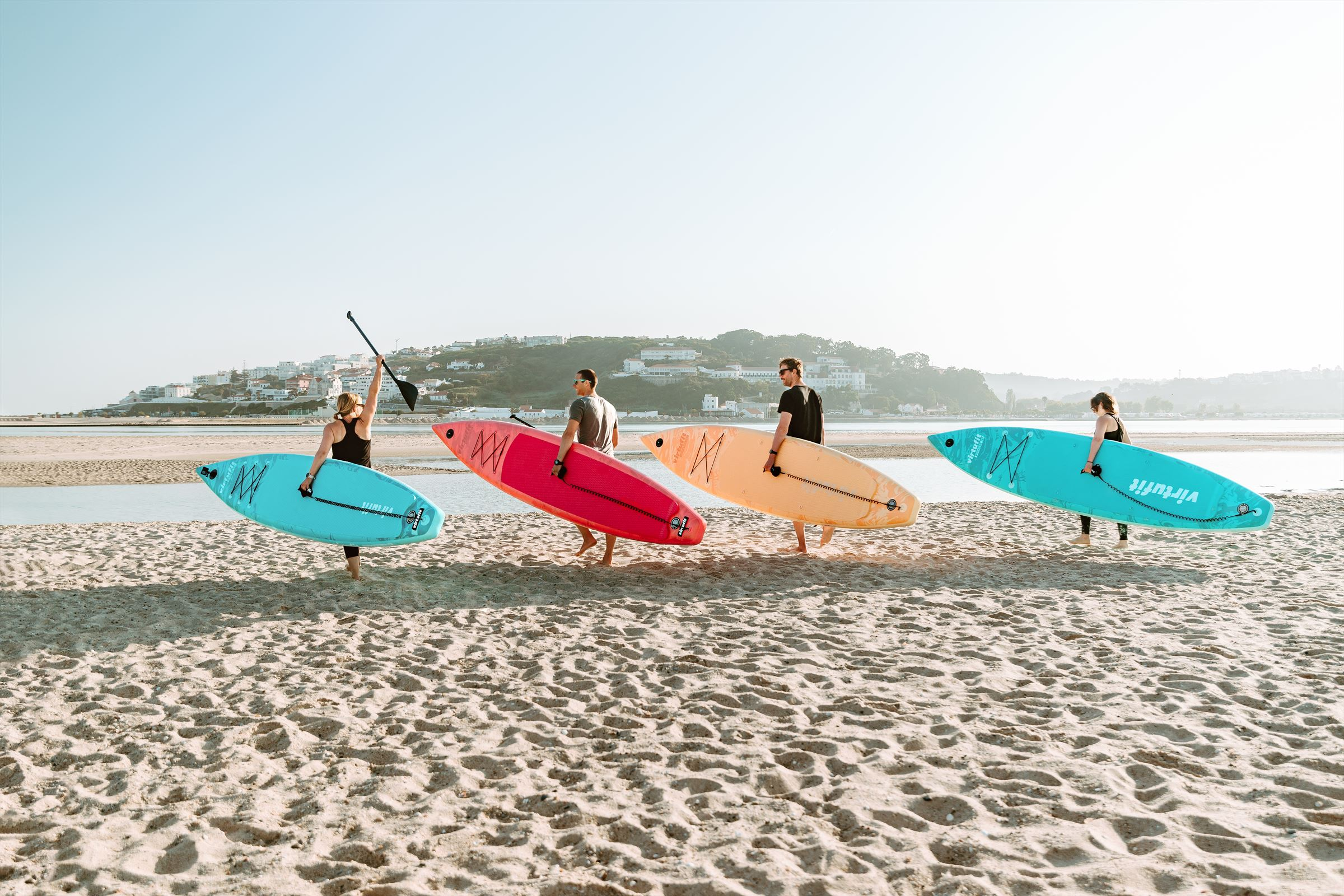 Menschen mit Paddleboards am Strand. Sie gehen auf das Wasser zu. Gebäude sind im Hintergrund.