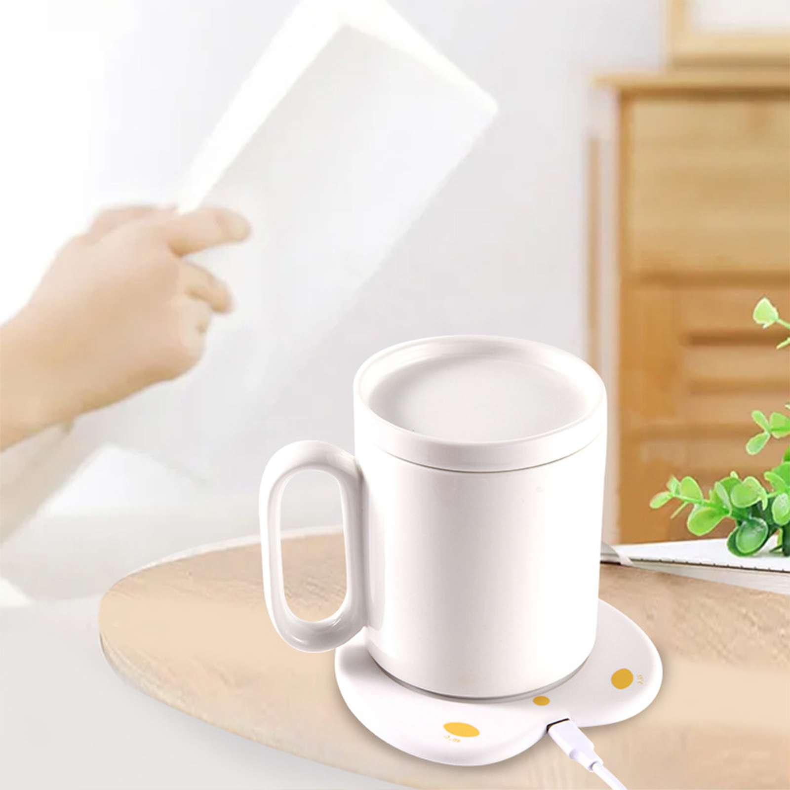 Eine weiße Tasse mit Deckel steht auf einem weißen Wärmer. Eine Hand hält im Hintergrund ein Buch. Eine grüne Pflanze und ein Schrank sind sichtbar.
