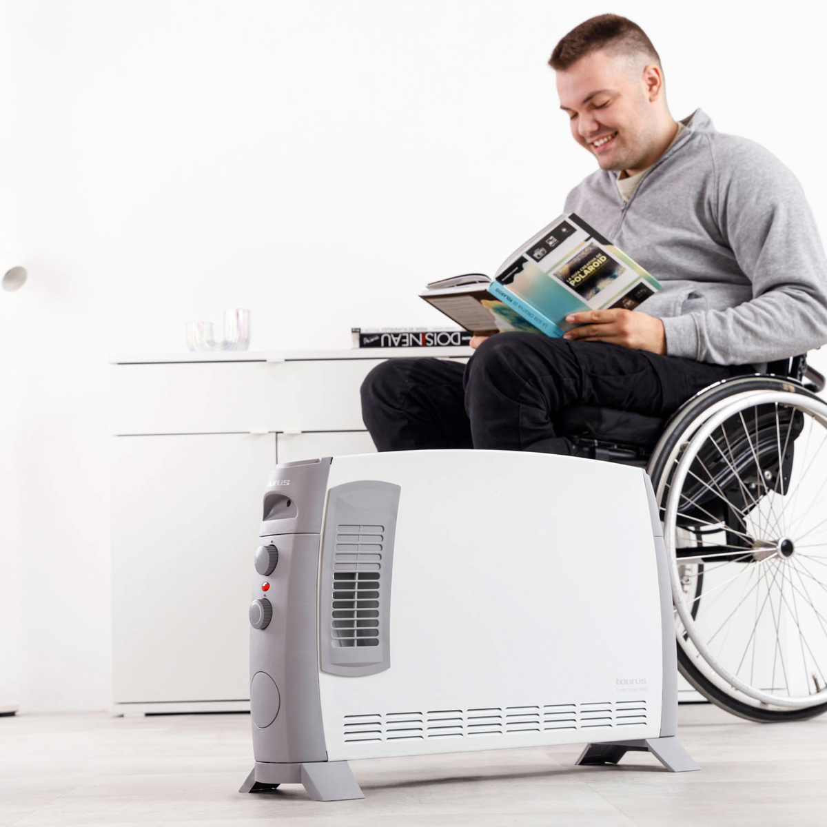 Un uomo in sedia a rotelle legge un libro. Un termoventilatore bianco è di fronte a lui. Un armadio bianco è alla sua sinistra.