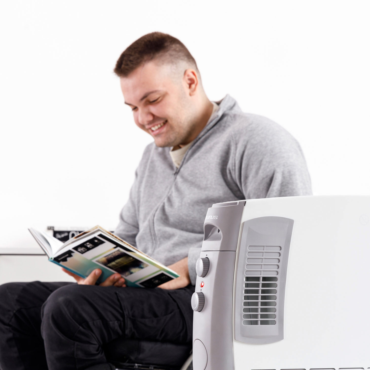 Un uomo sorride leggendo un libro, un termoventilatore bianco è in primo piano.