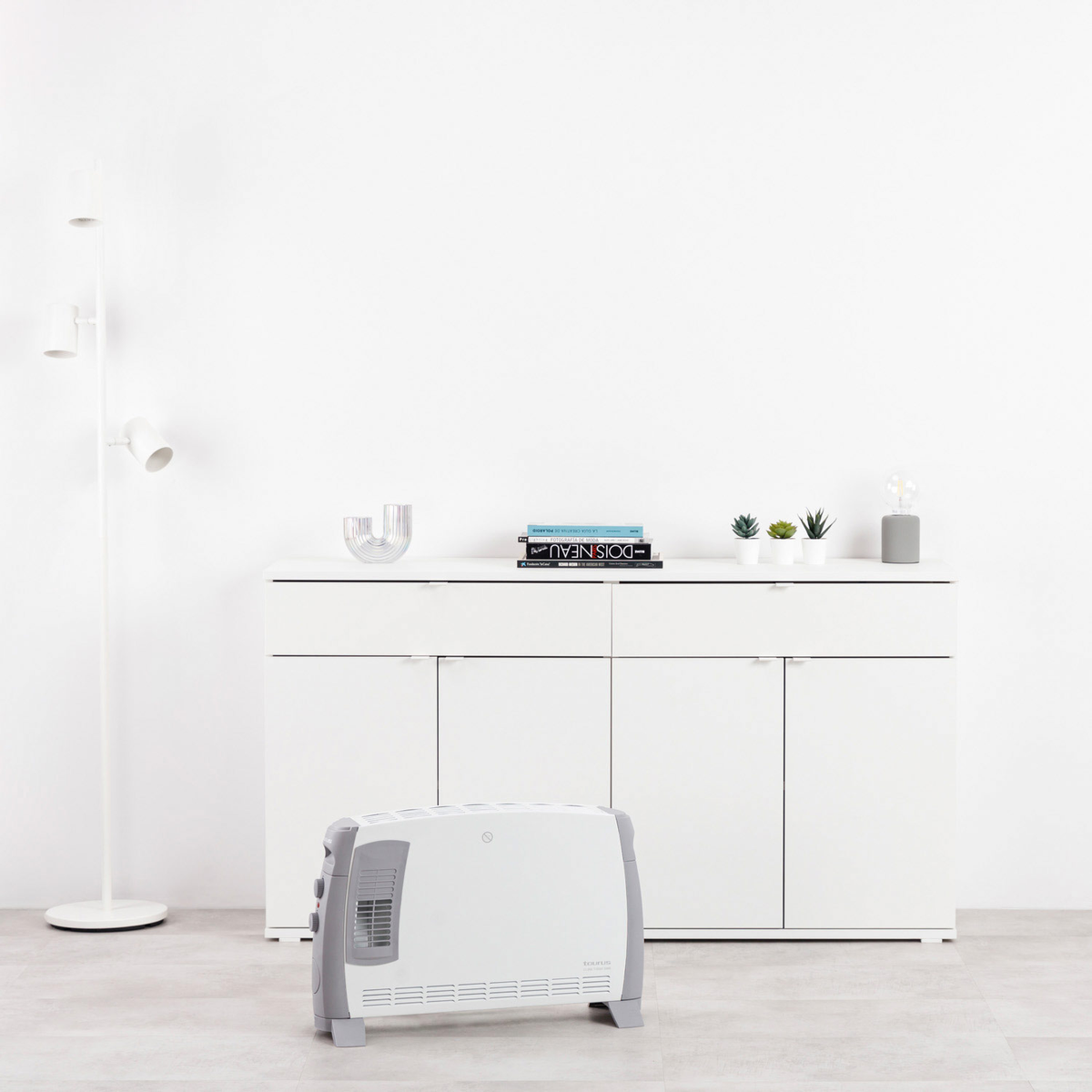 Un termoconvettore elettrico bianco e grigio è appoggiato a terra davanti a un mobile bianco con libri e piante. Una lampada da terra a sinistra.