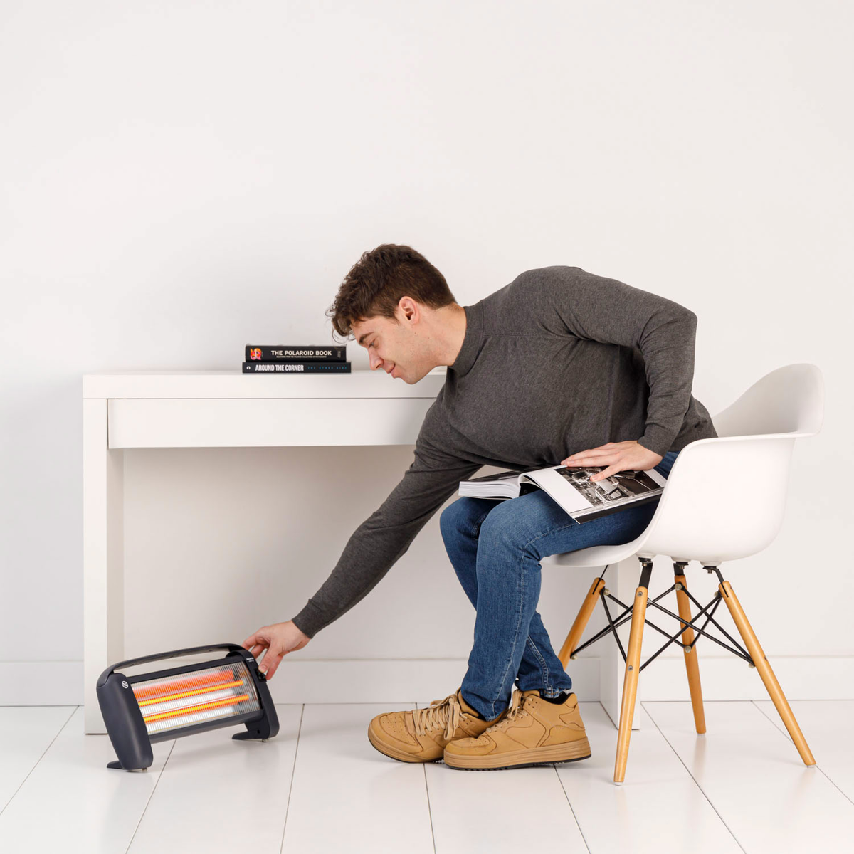 Un uomo in jeans e camicia grigia è seduto e legge. Tocca un riscaldatore nero e arancione. C'è una scrivania bianca con libri e una sedia bianca.