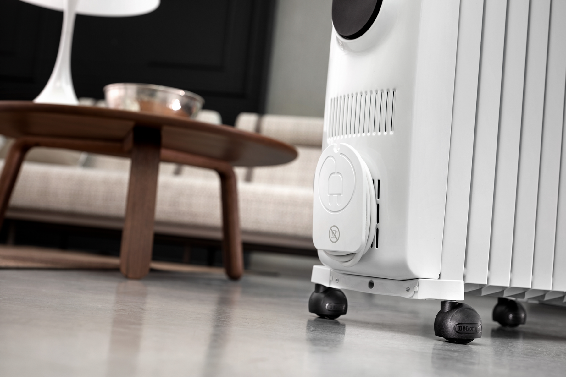 Biały grzejnik olejowy na szarej podłodze. Drewniany stół i sofa są rozmyte w tle.