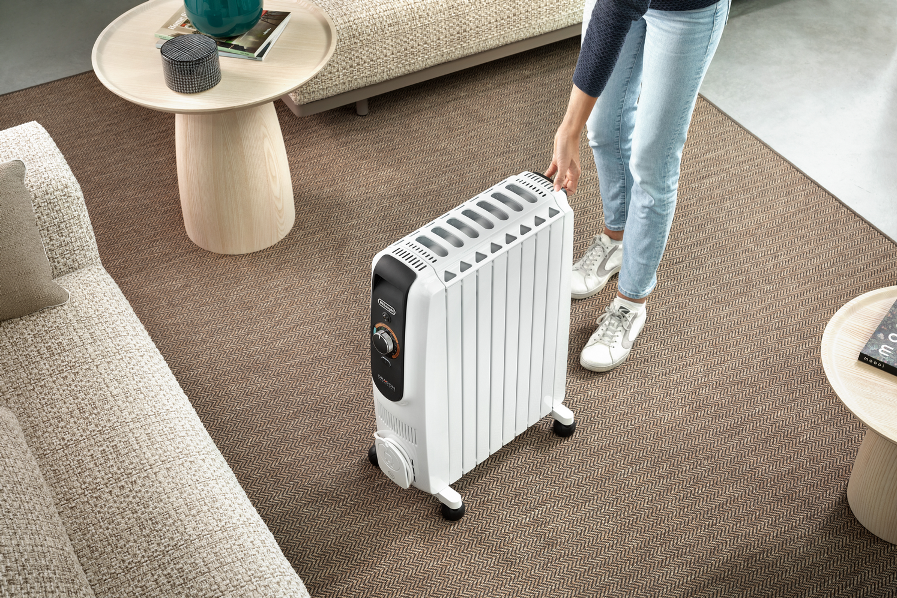 Osoba przesuwa biały kaloryfer olejowy po brązowym dywanie. W pokoju znajduje się sofa i stoliki boczne.