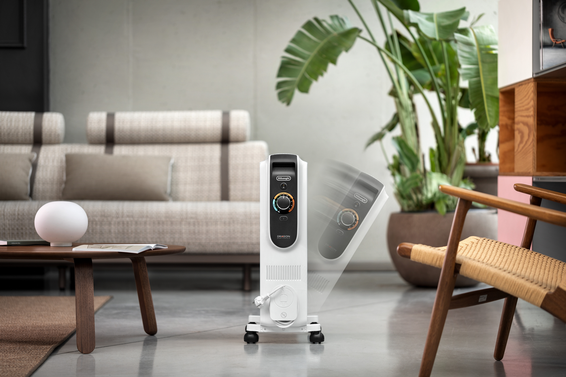 Biały grzejnik DeLonghi Dragon w pokoju. Obecne są kanapa, roślina, stół i krzesło. Widoczny panel sterowania i przewód grzejnika.