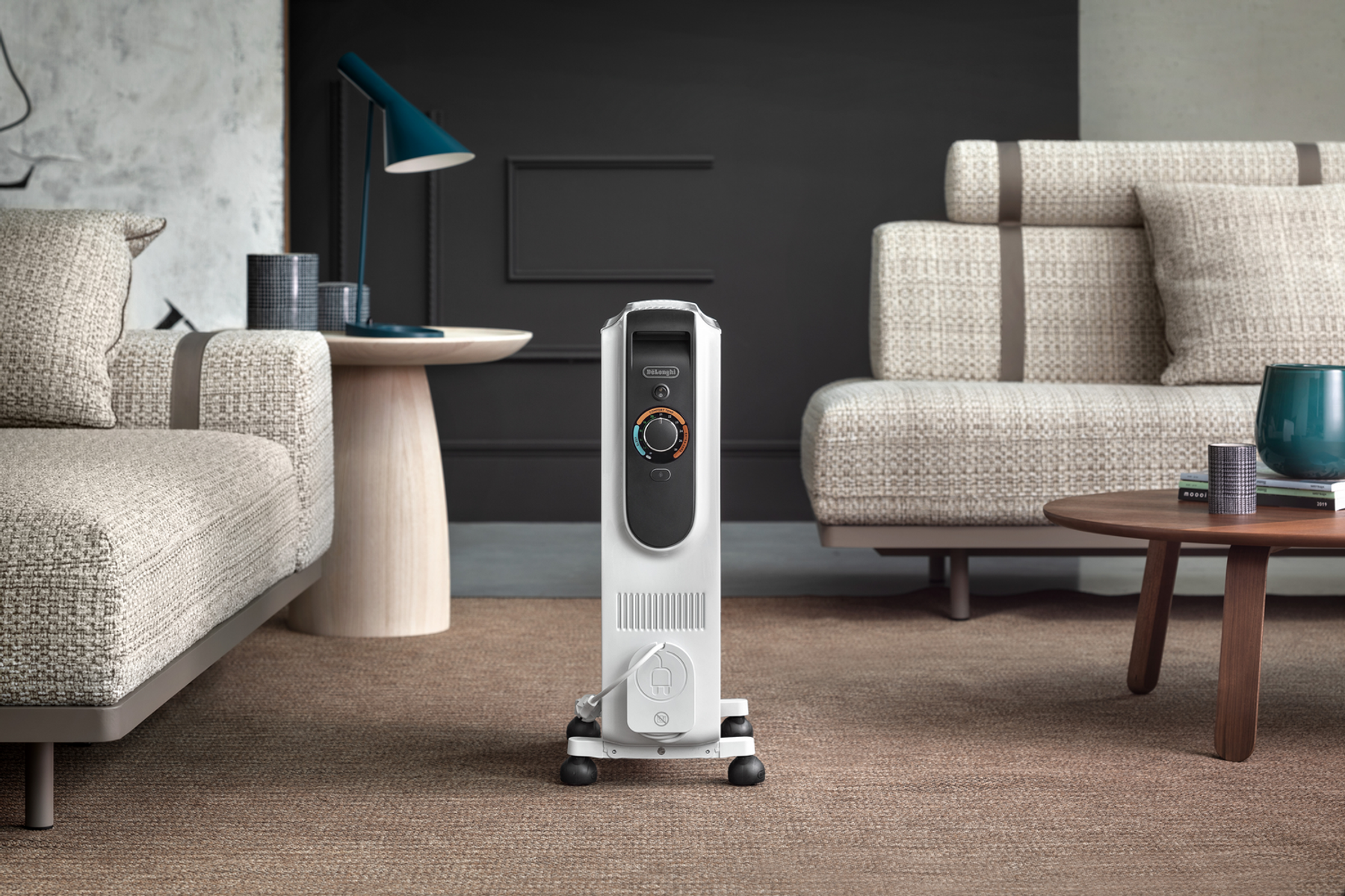 Biały grzejnik olejowy stoi na dywanie. W tle sofa, stół i lampa. Ściana jest ciemna.